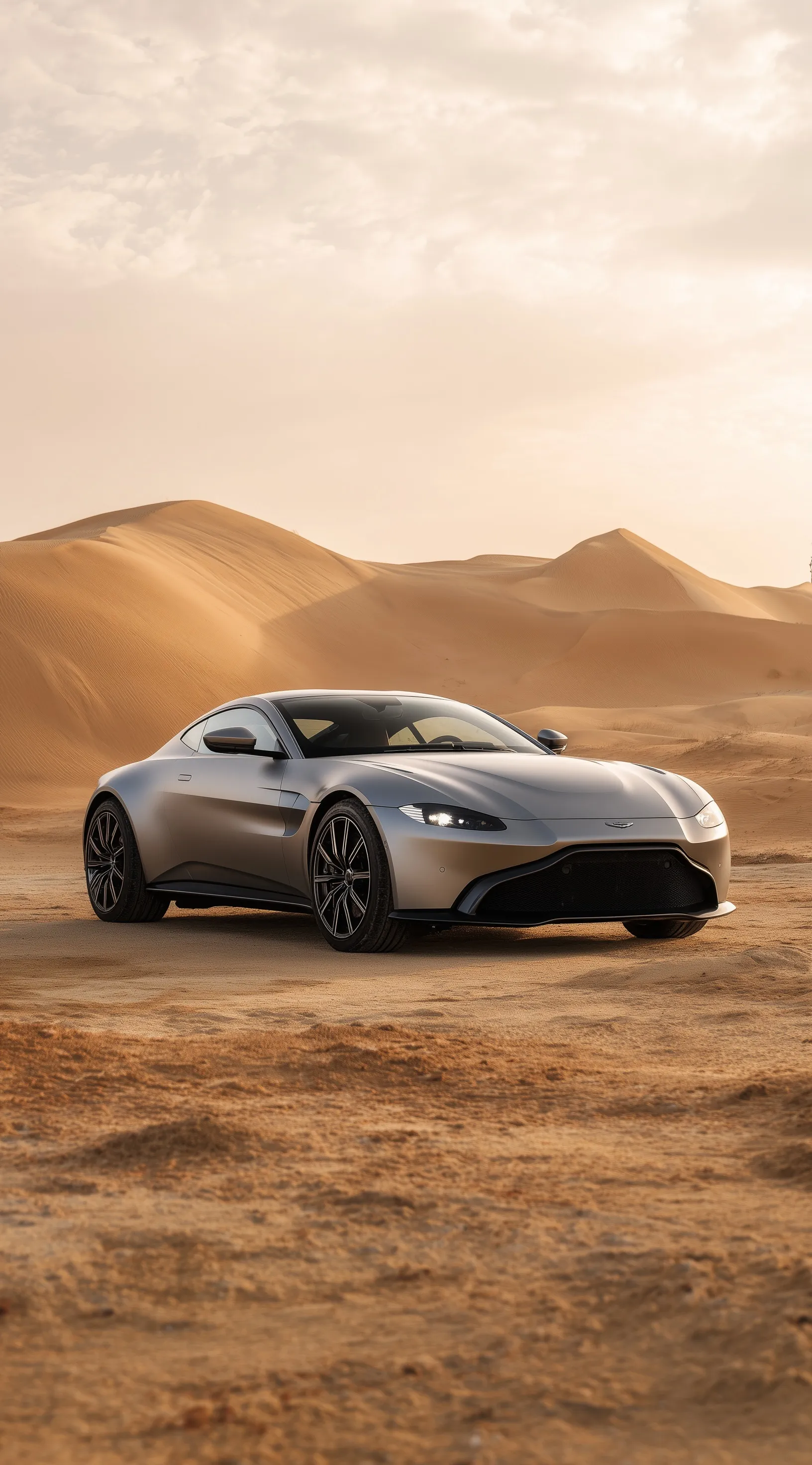 A sleek light grey Aston Martin V8 concept car parked on desert dunes, perfect for iPhone wallpaper backgrounds.