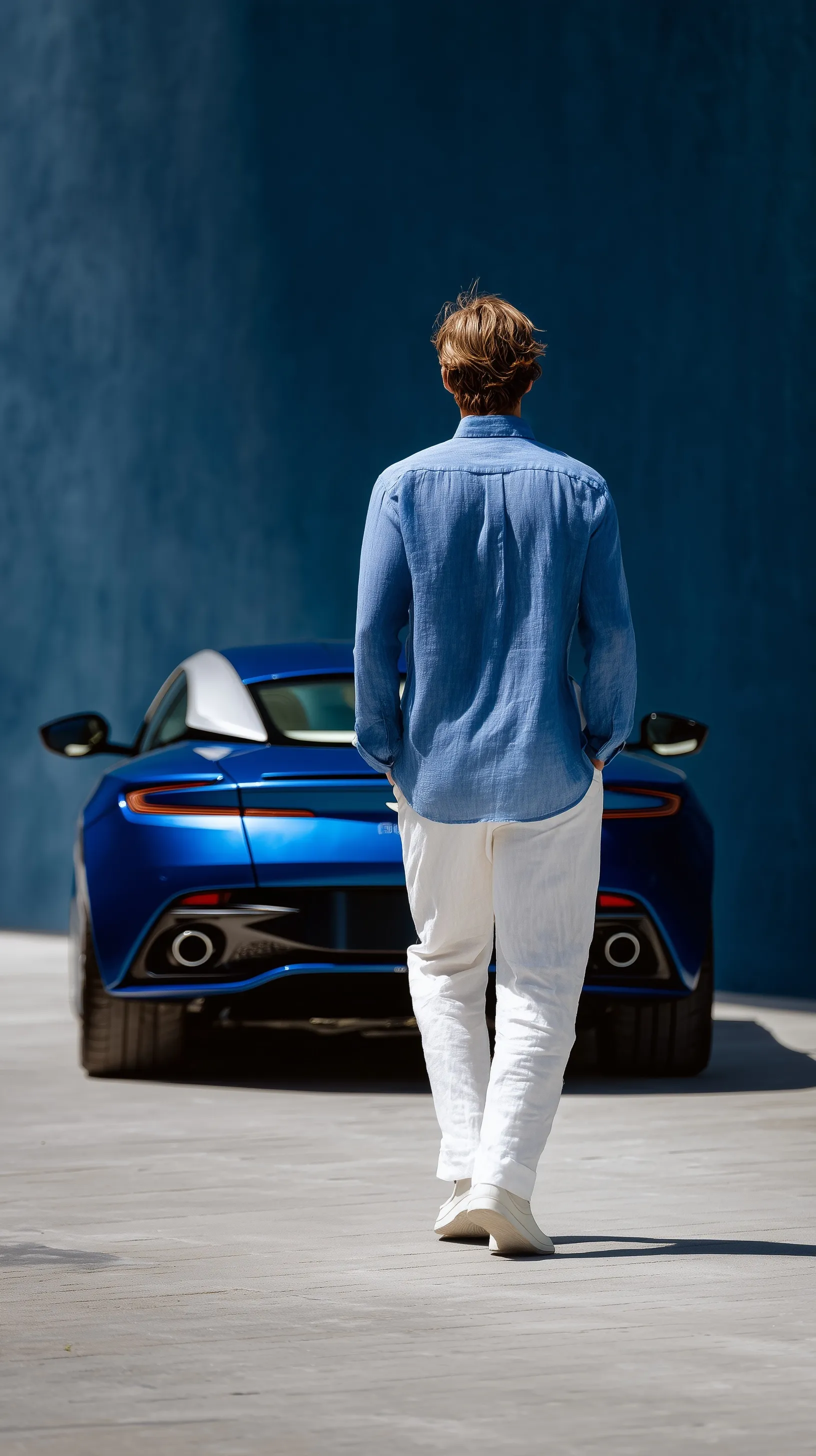 Elegant man in linen shirt walking toward a blue Aston Martin DB12 against a dark grey aesthetic wall.