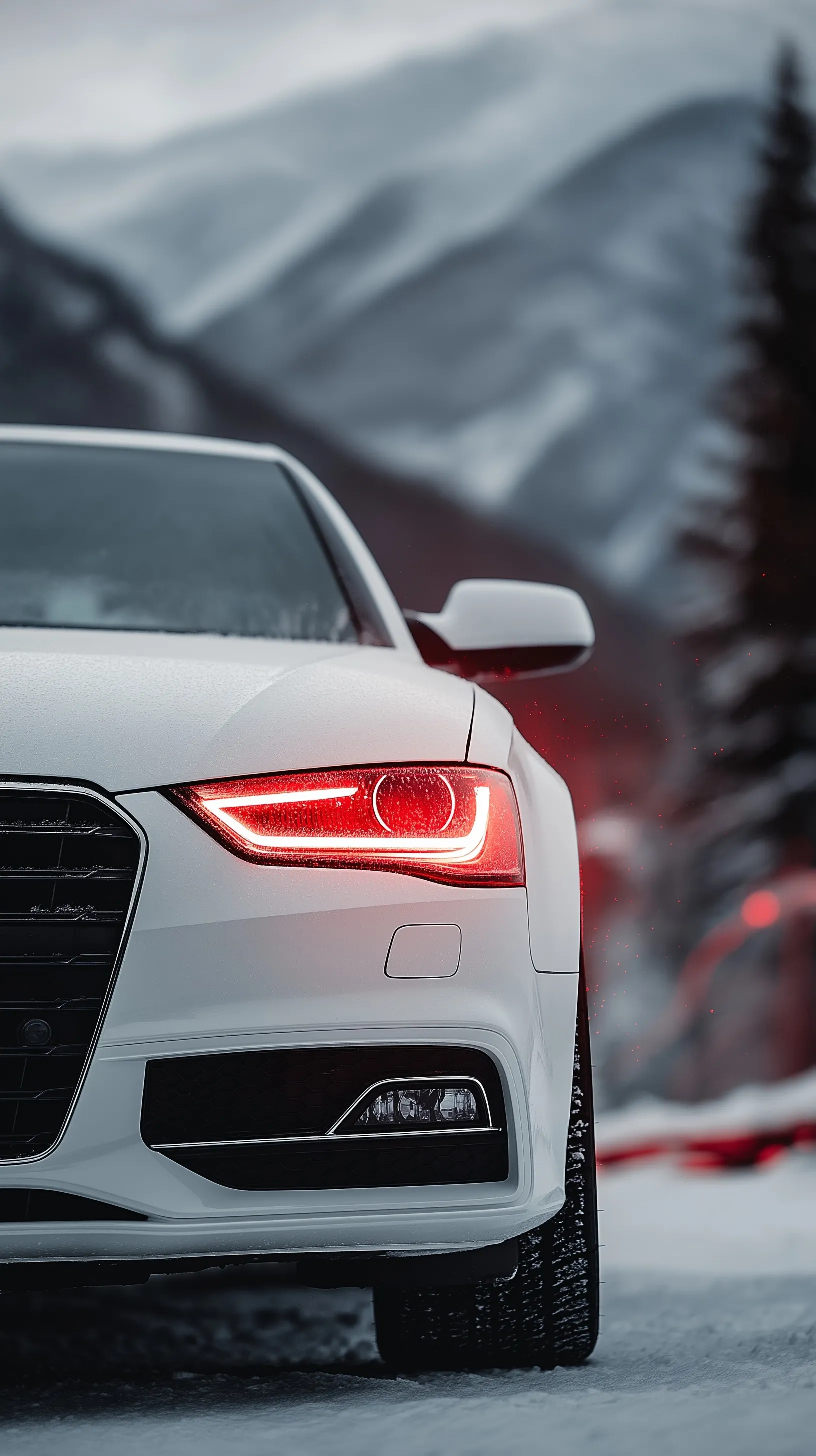 Front view of a white Audi car with glowing red headlights parked against a blurred snowy mountain landscape.