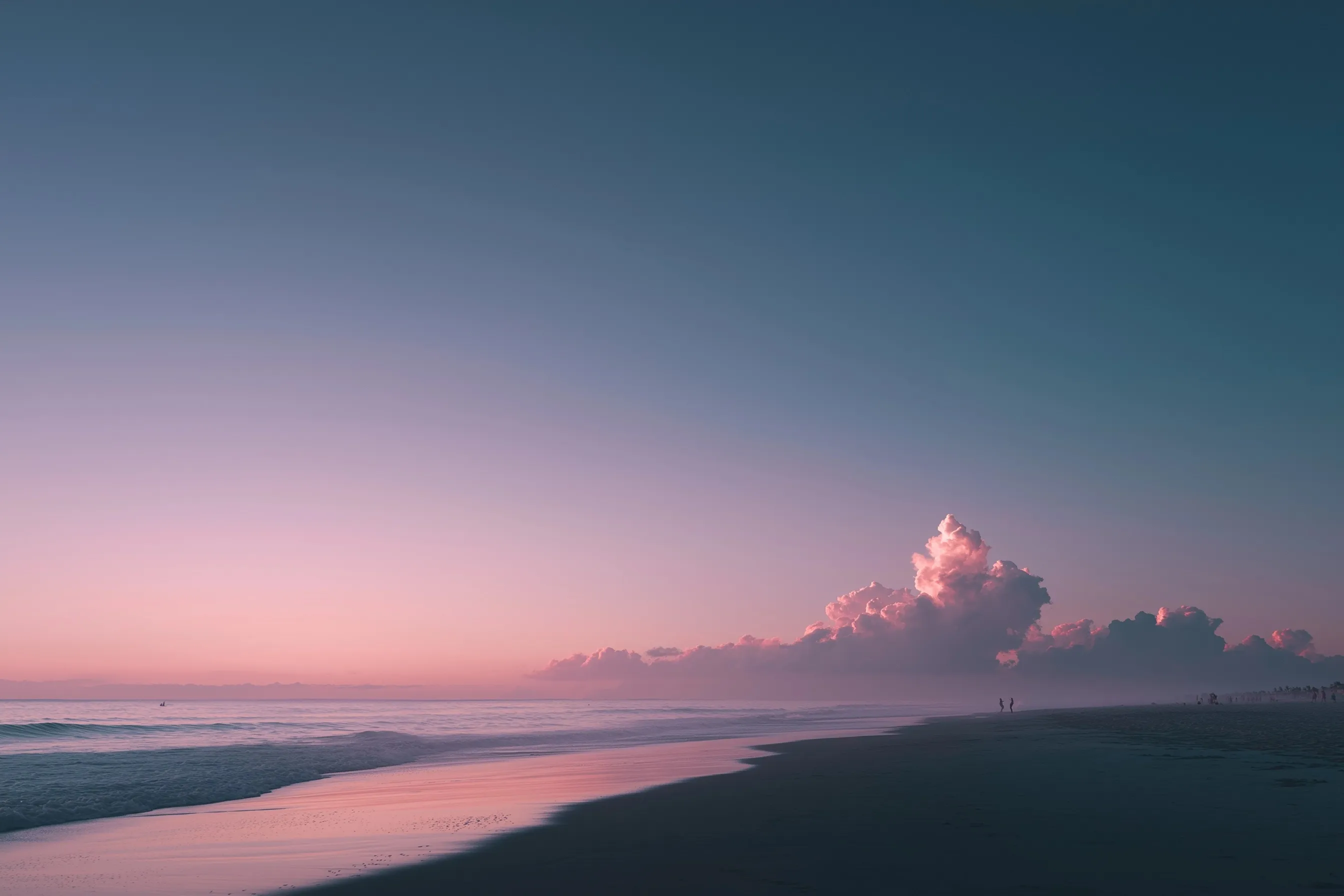Tropical beach sunset with a coral to indigo gradient sky, soft clouds, and distant surfer silhouettes.