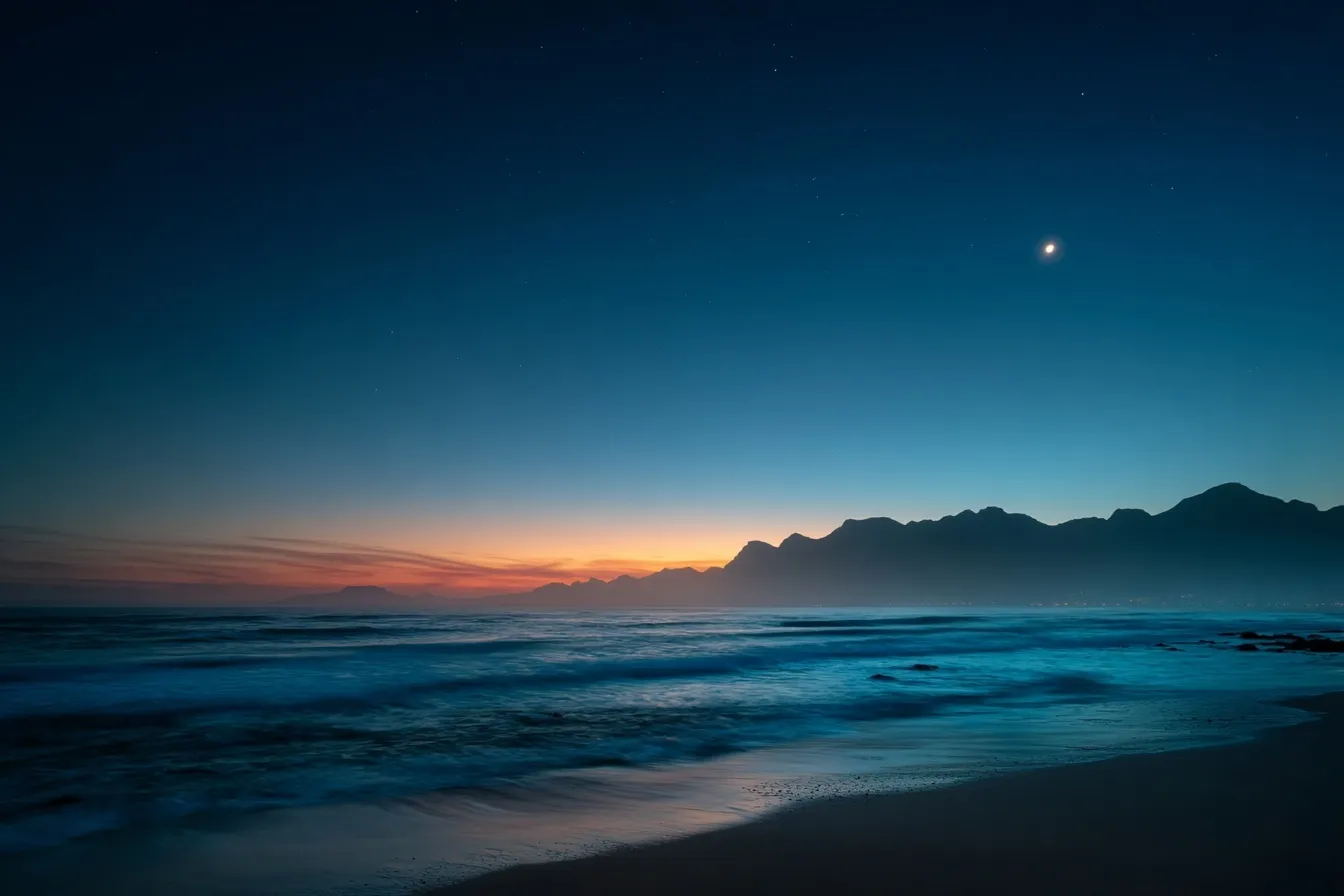 Long exposure beach sunset with mountains, dark blue sky, orange horizon streaks, and a crescent moon.