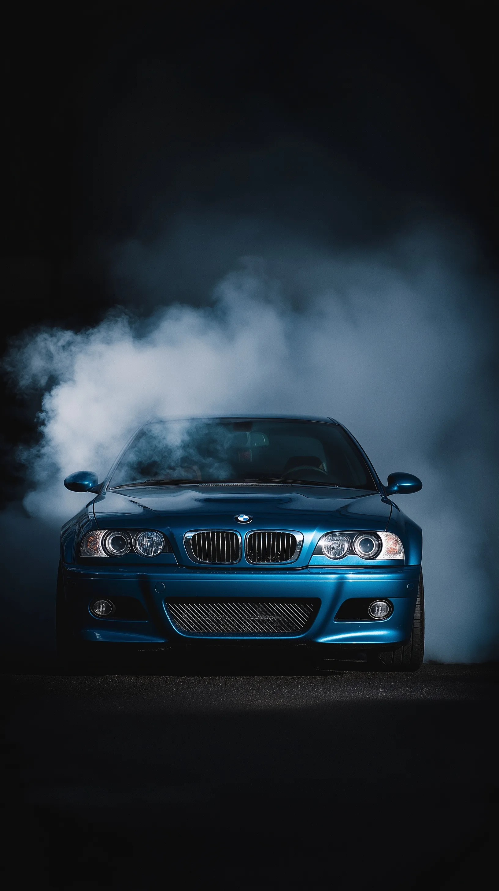 Blue BMW E46 M3 front view with chrome grills surrounded by cinematic tire smoke against a dark moody background.