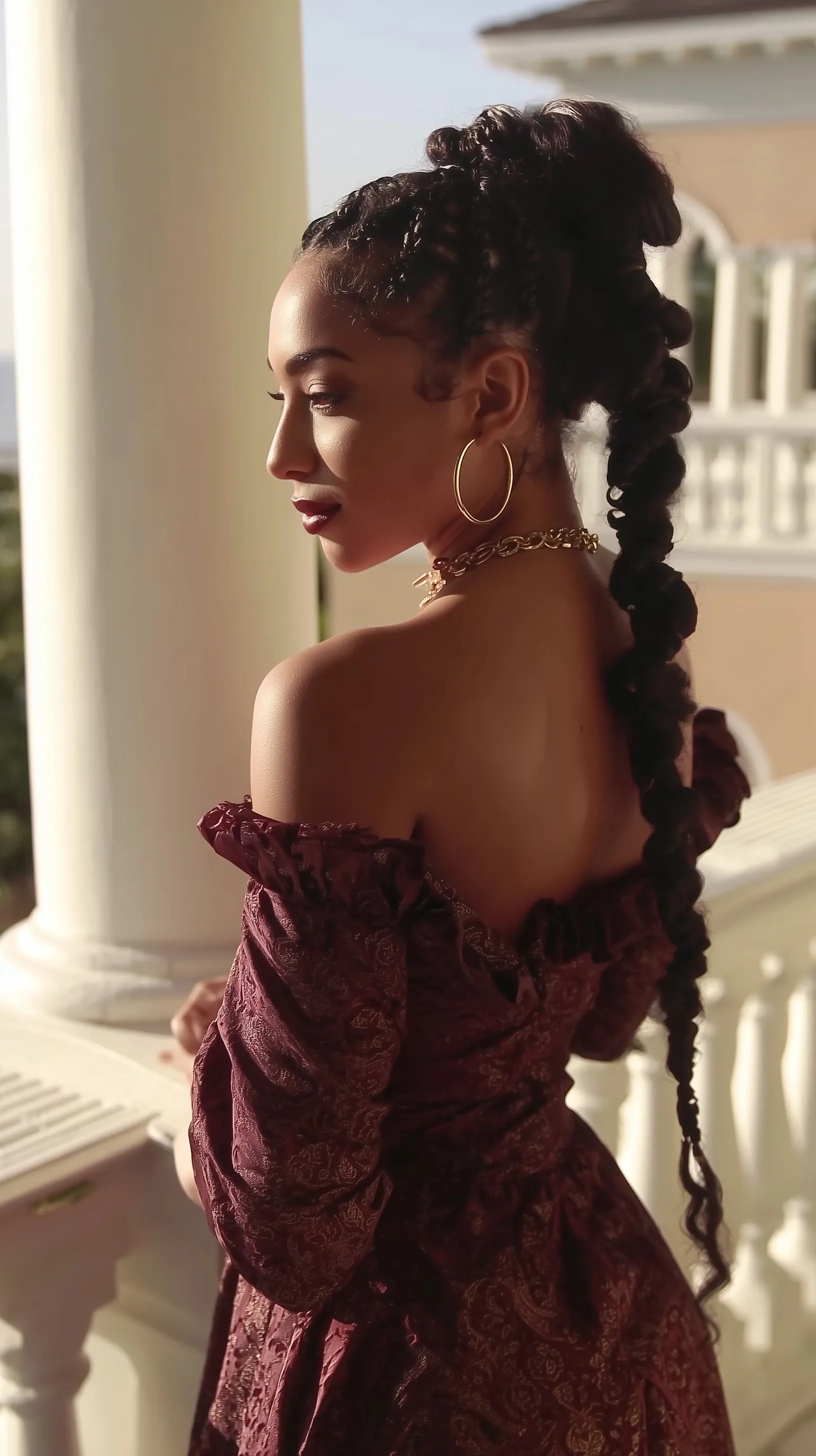 Exotic woman with long black braids and soft curls wearing a maroon dress, posing on a white balcony in profile.