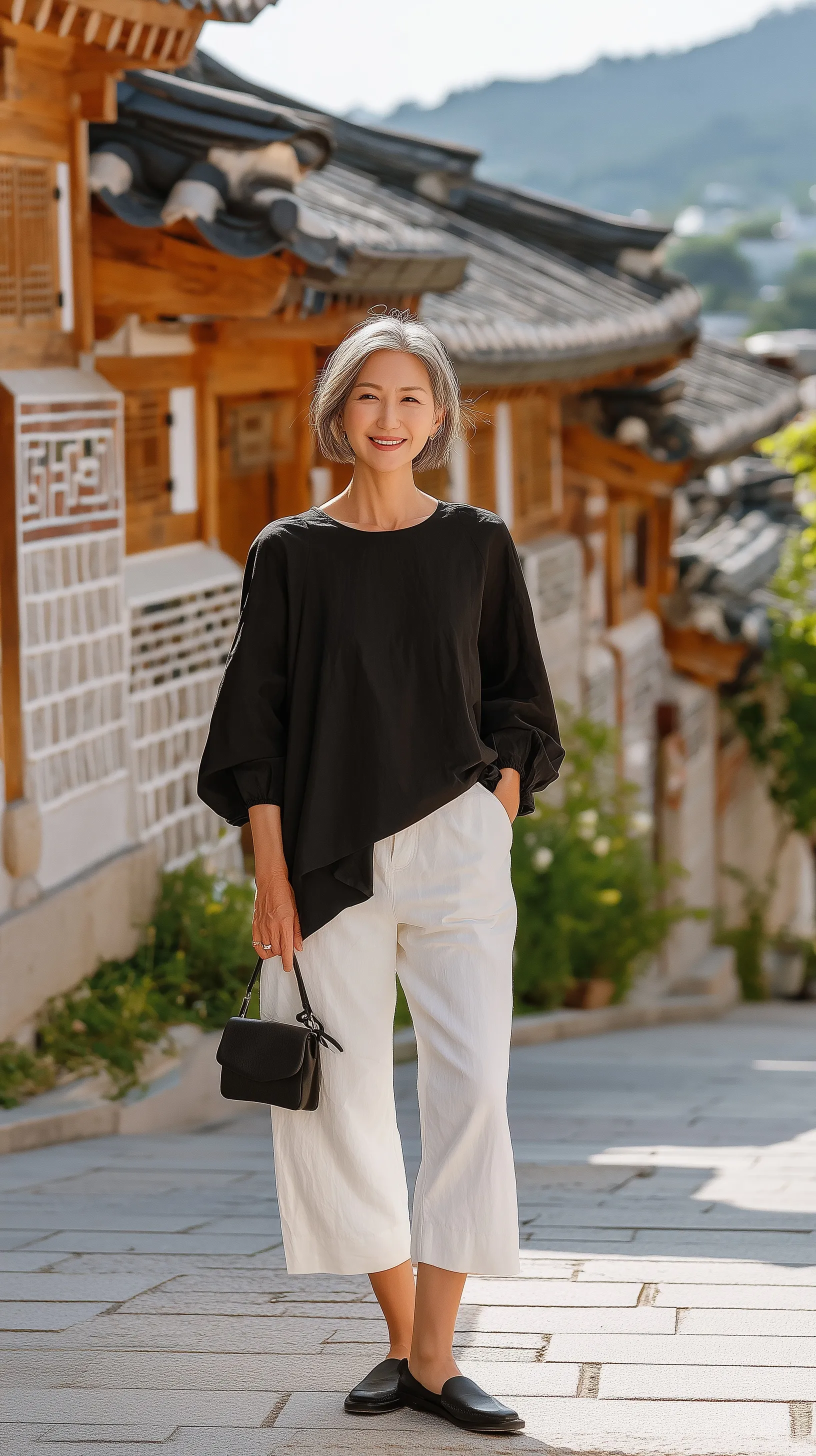 Elegant 60-year-old woman in a black pleated top and white capri pants posing in a traditional hanok alley.