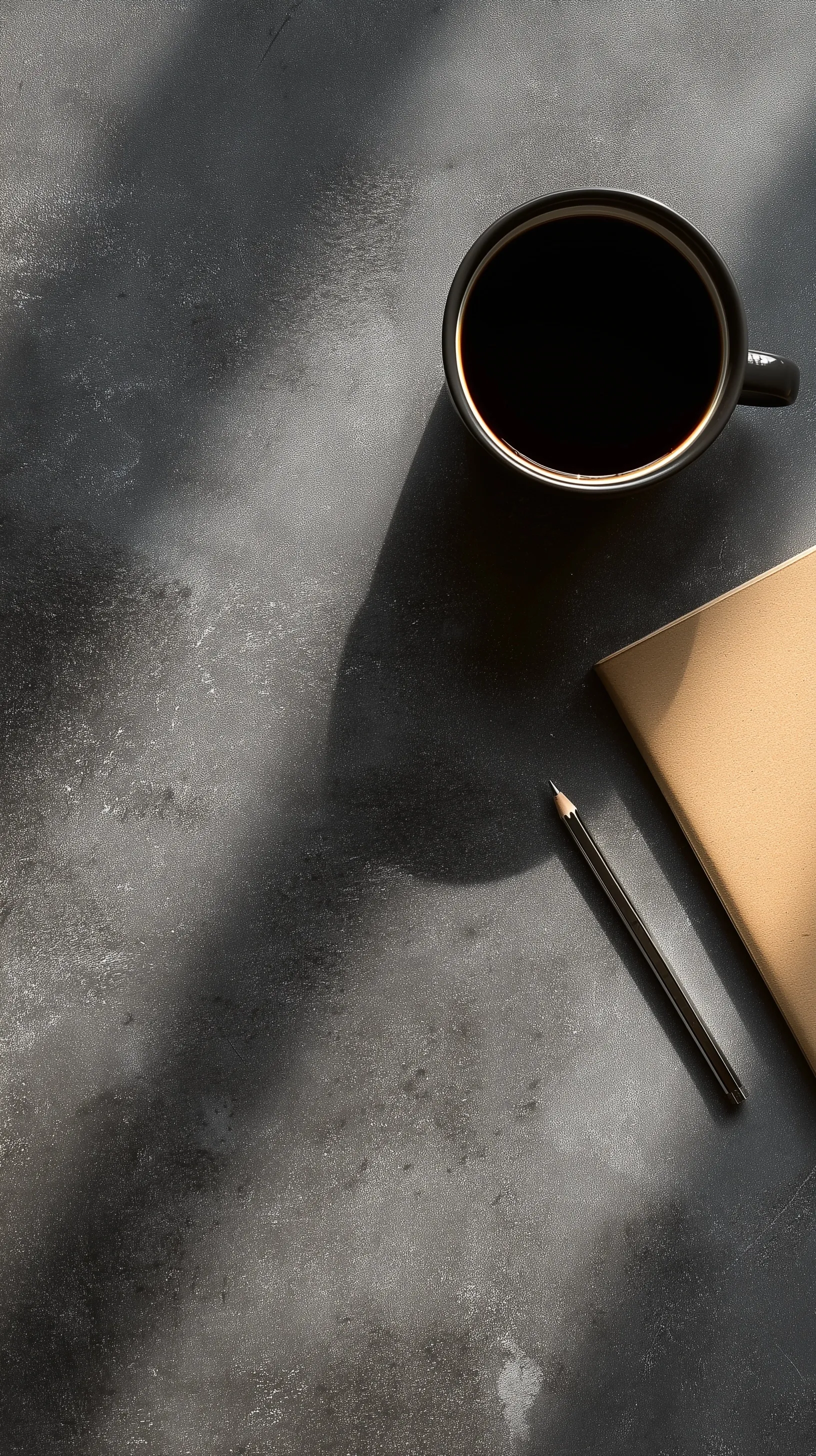 Flatlay of a black coffee mug, notebook, and pencil on a dark concrete industrial office desk with hard shadows.