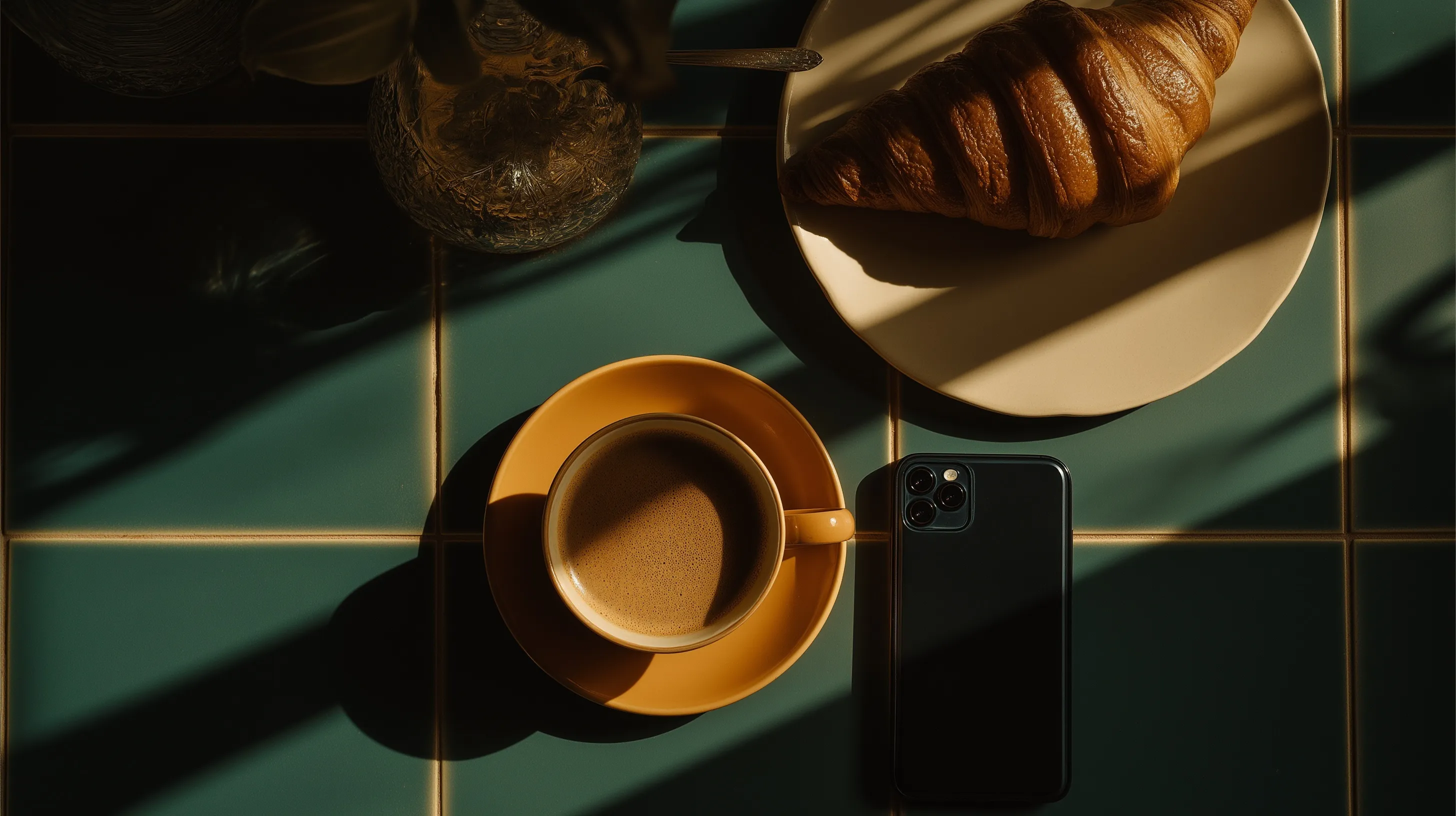 Top-down view of coffee, croissant, and smartphone on a green tiled table with warm morning sunlight.