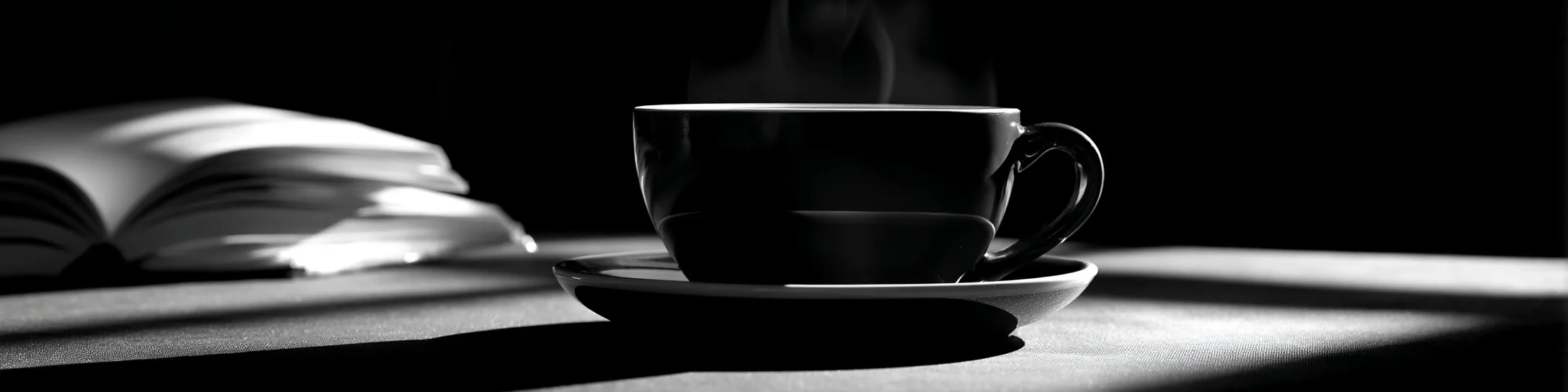 A black and white close-up of a coffee cup on a table with dramatic rim lighting, steam, and scattered books.