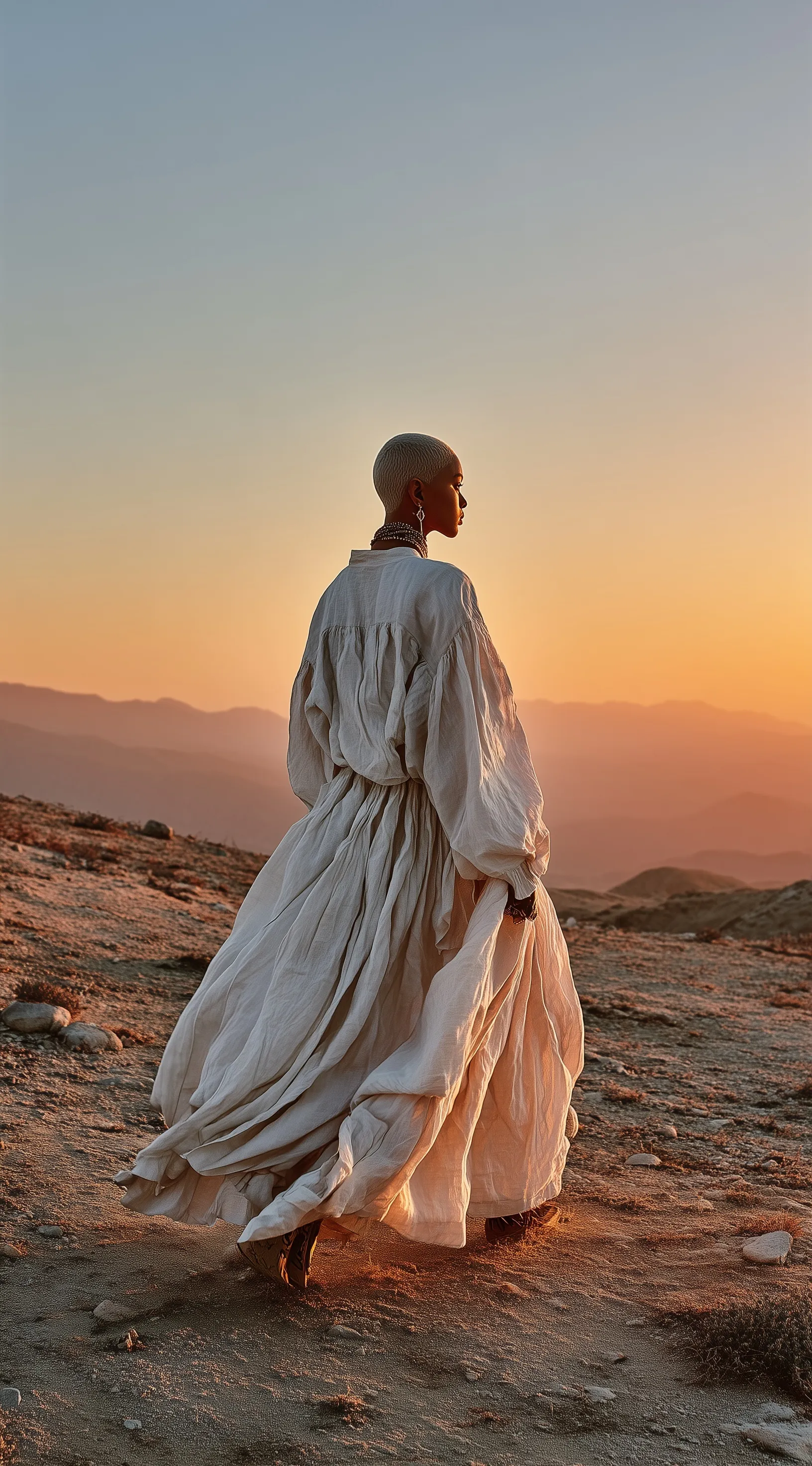 A woman with buzzed hair in a white linen dress walking through a desert landscape at dusk.