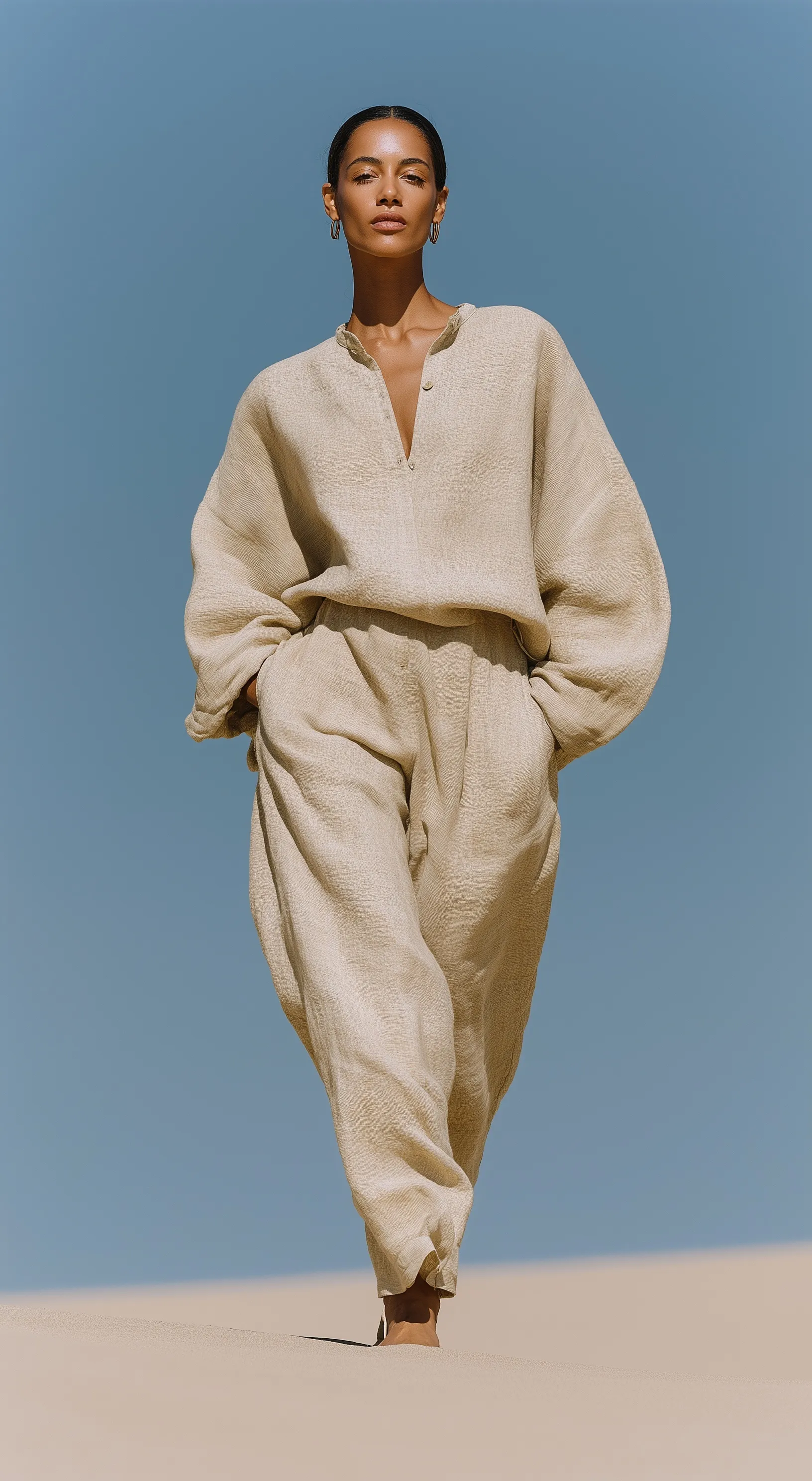 Arab woman in flowing sand-colored linen outfit standing on a desert dune ridge during golden hour.