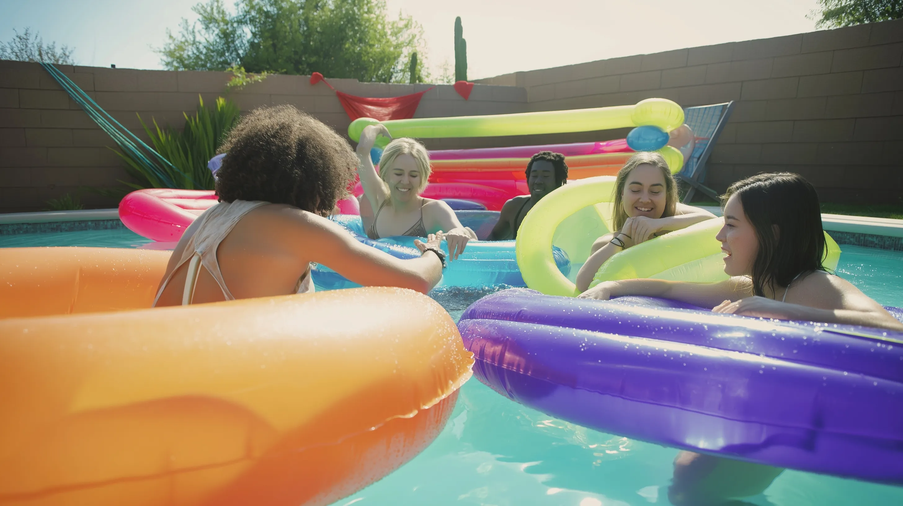 A group of friends at a neon pool party with colorful floaties, captured in an authentic, candid handheld style.