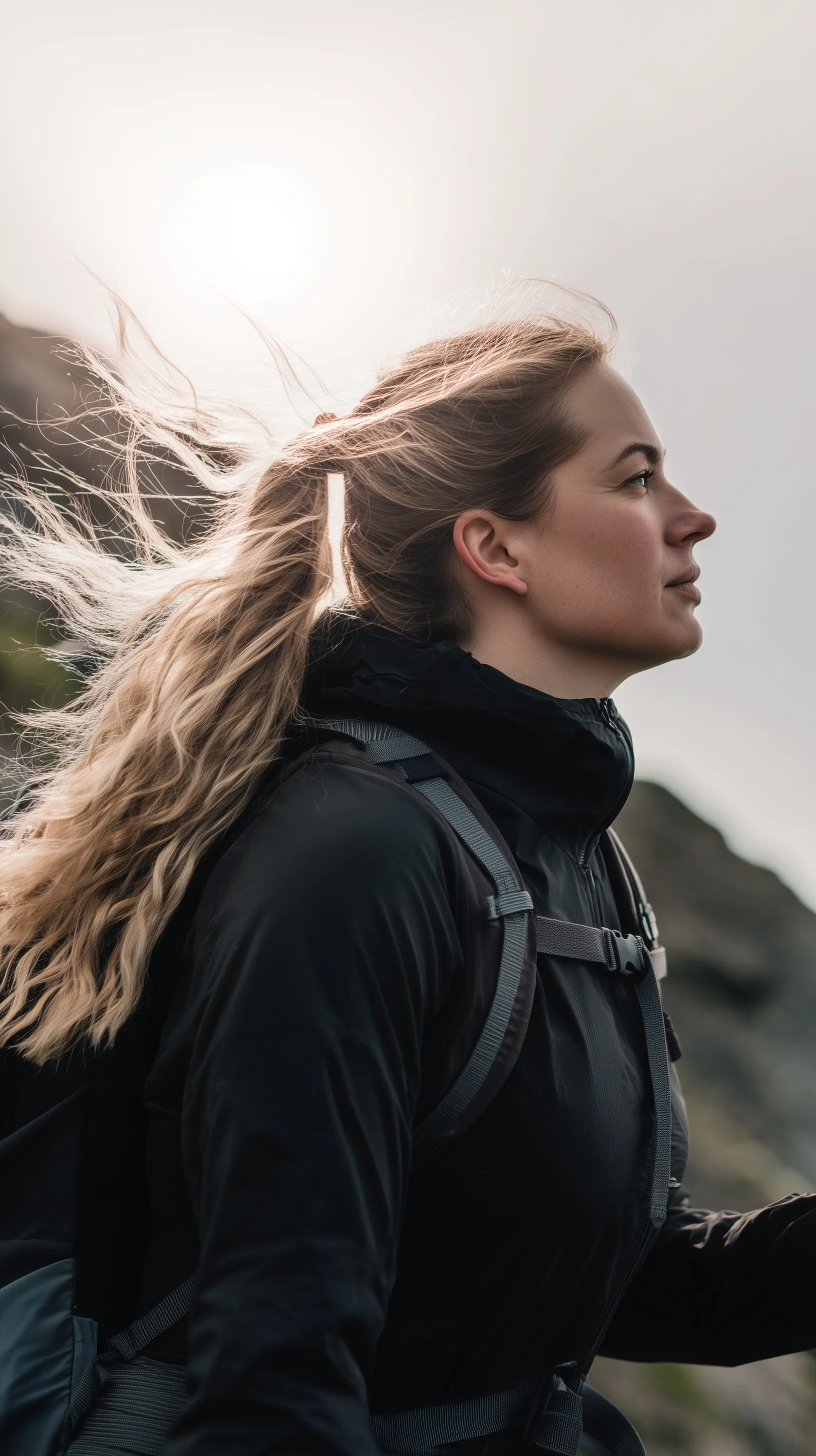 A female hiking guide in modern techwear posing outdoors in strong wind, showcasing functional mountain apparel.