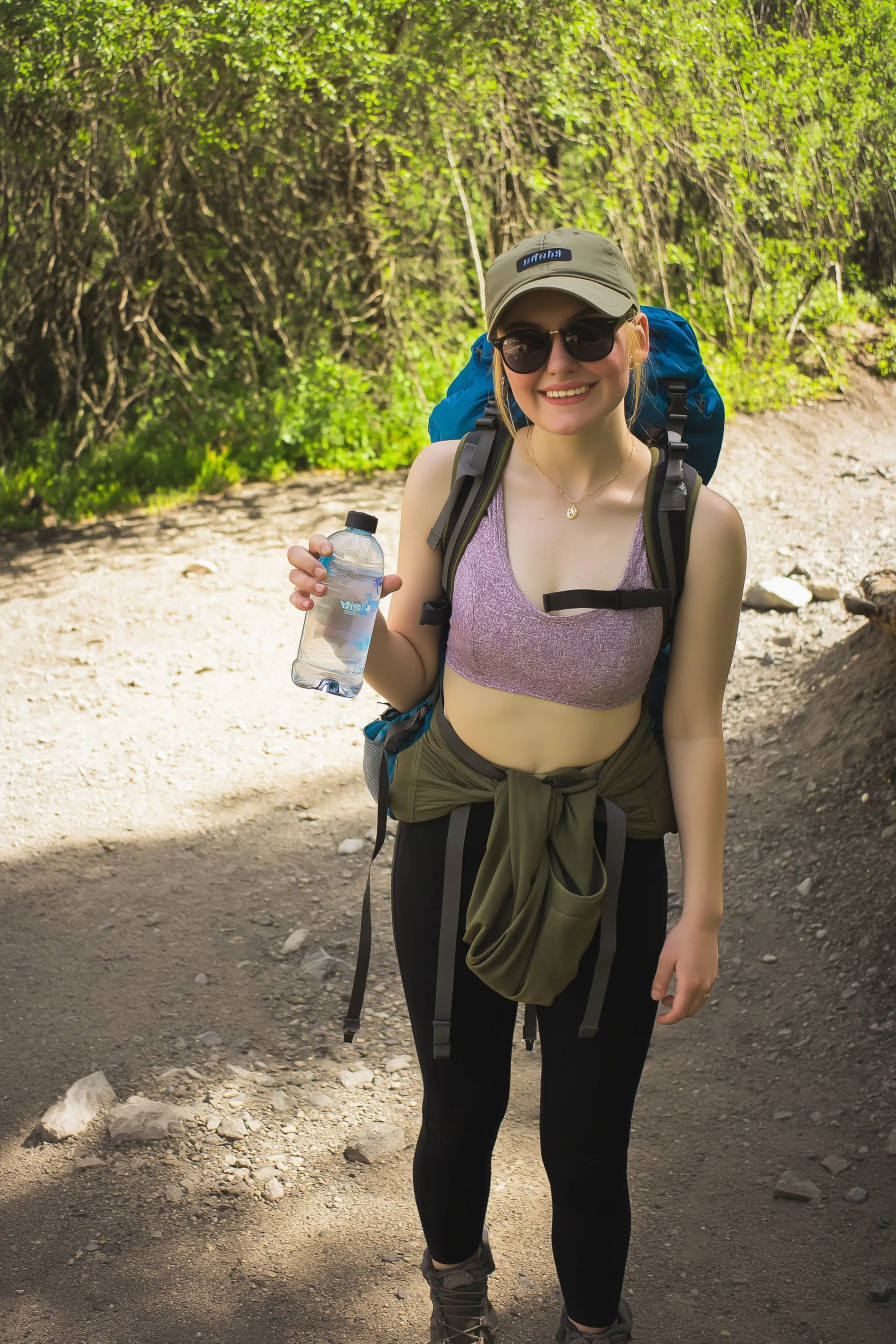 A young woman with a hiking backpack and water bottle on a trail, wearing dirty hiking boots and a ball cap.