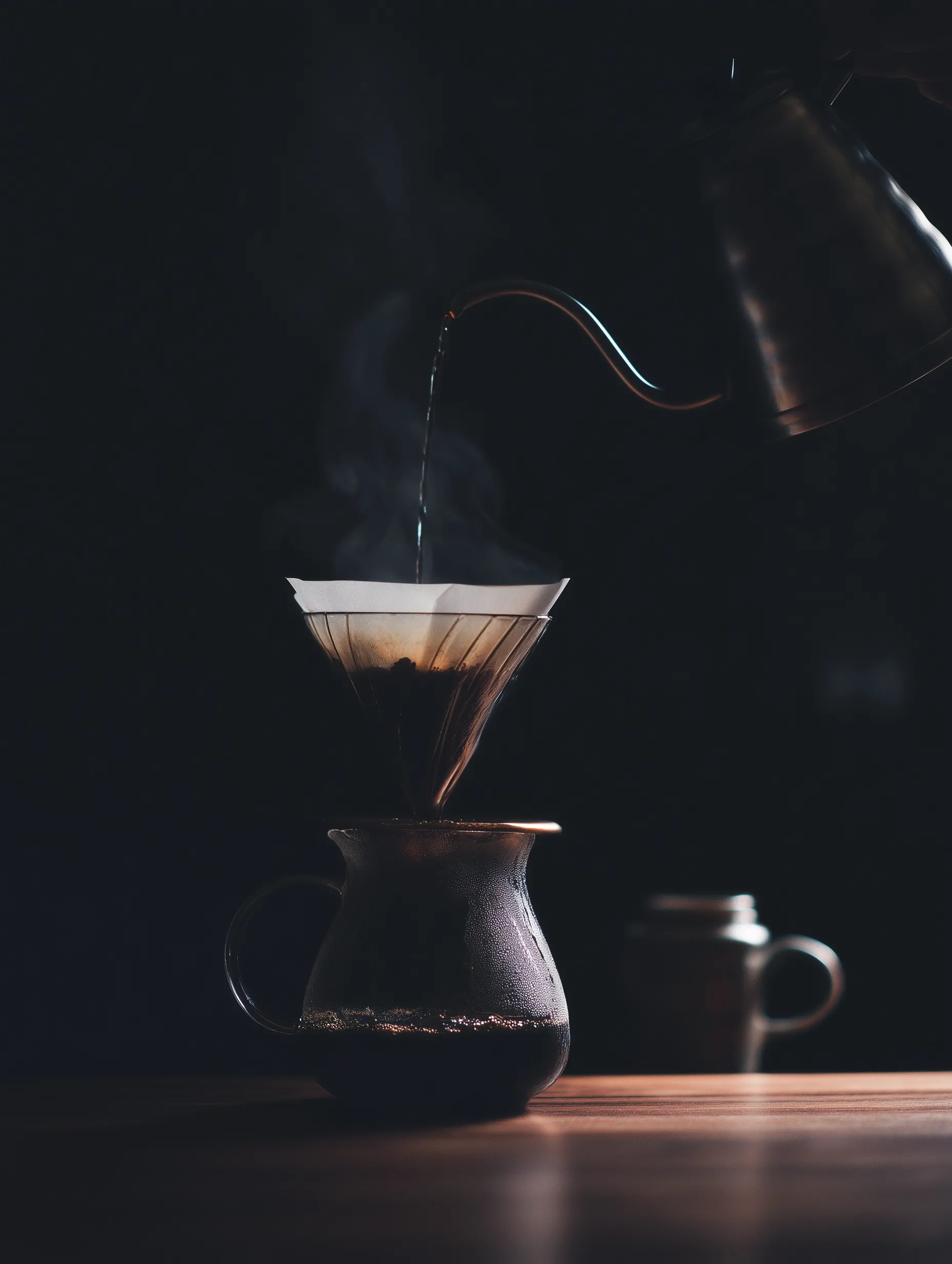 A dark, cinematic low-key shot of hot water being poured from a metal kettle into a coffee dripper.