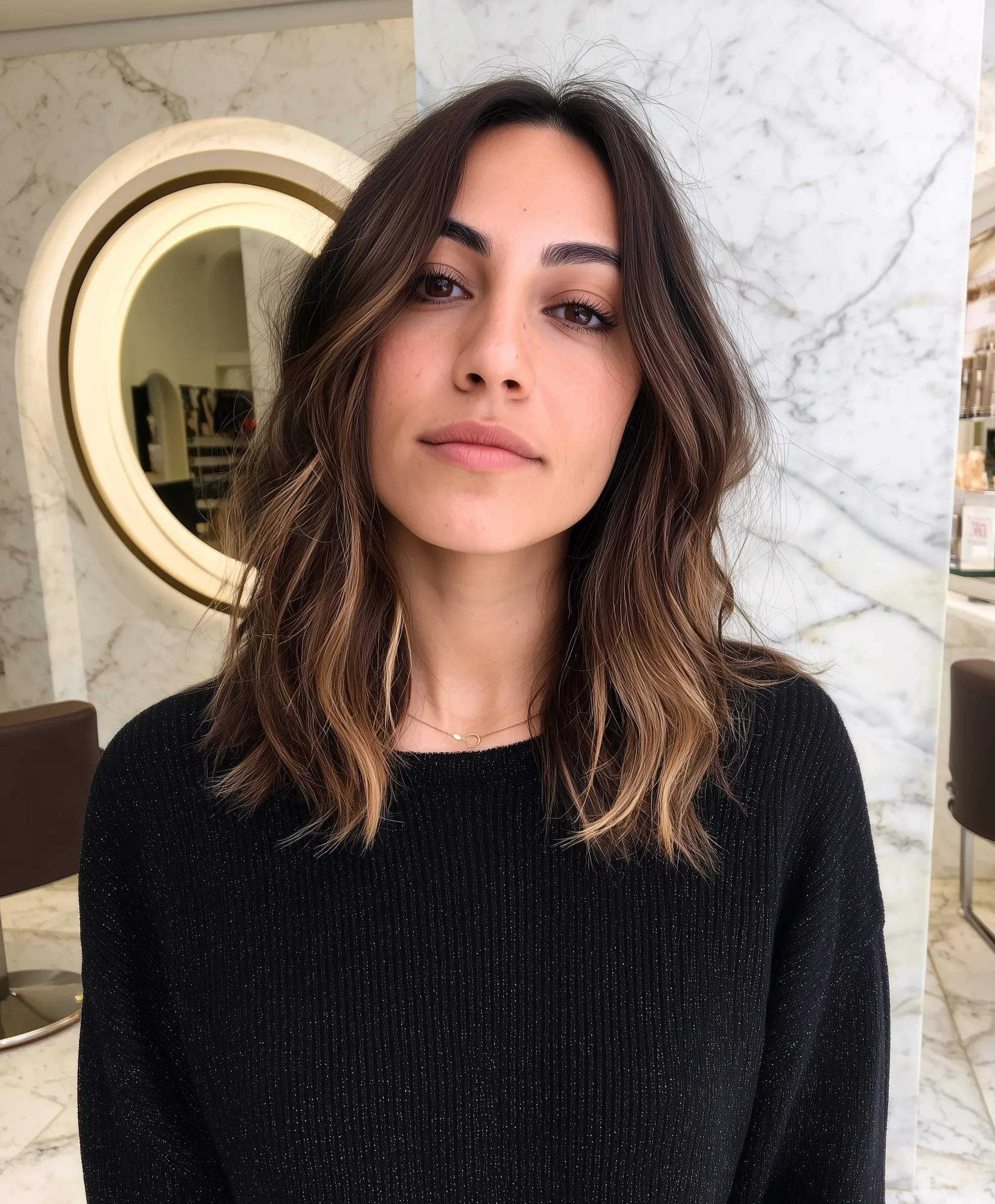 A 32-year-old brunette woman with a caramel balayage lob hairstyle posing in a modern salon with a circular mirror.