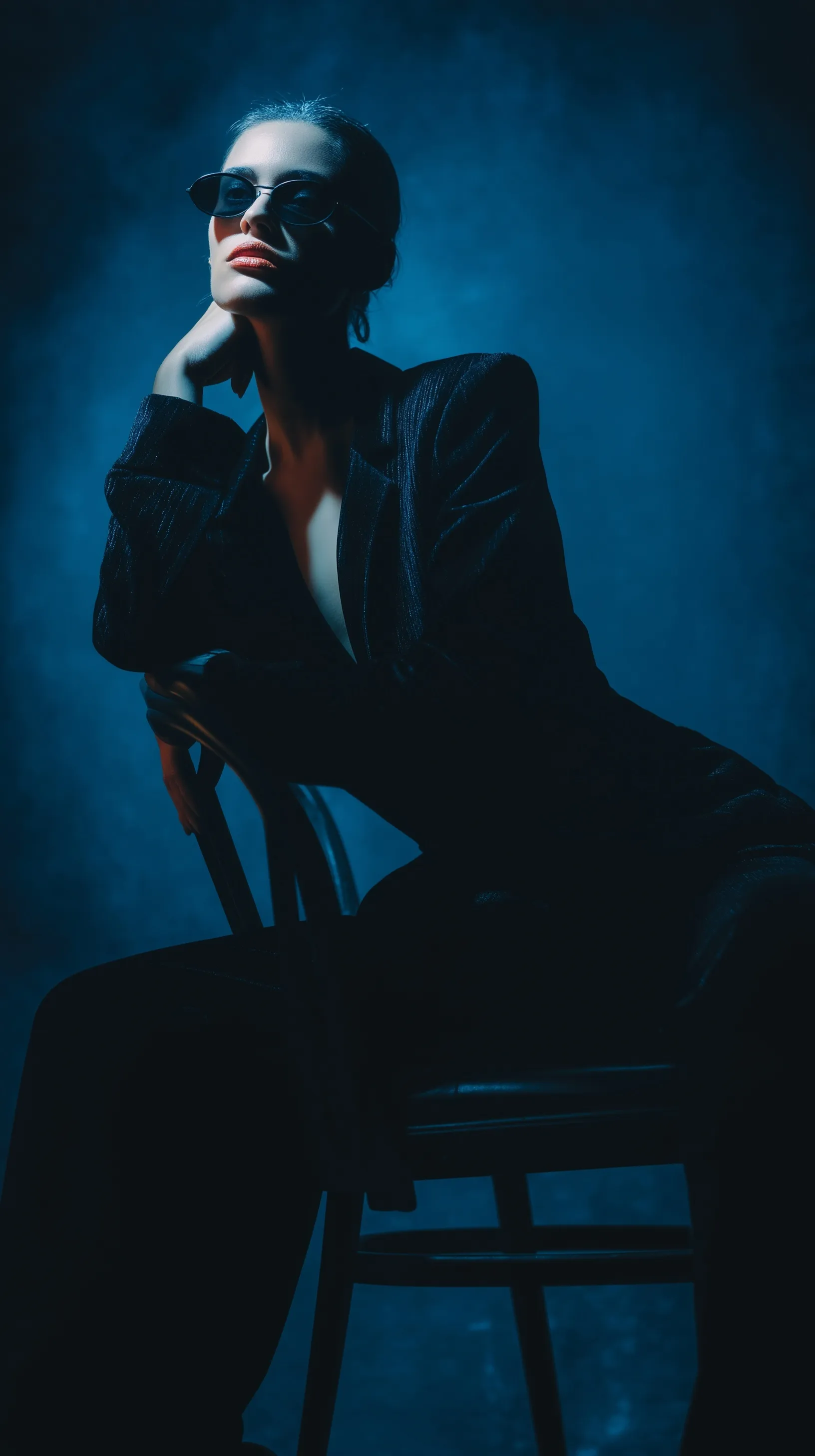 A moody fashion portrait of a stylish woman in a black outfit and sunglasses sitting on a chair in a dark club.