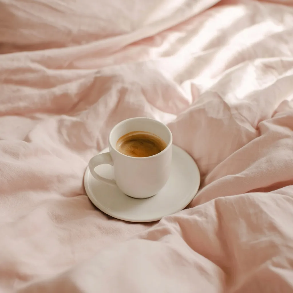 A white coffee cup on a pink saucer atop soft, crumpled bedding with gentle morning light.