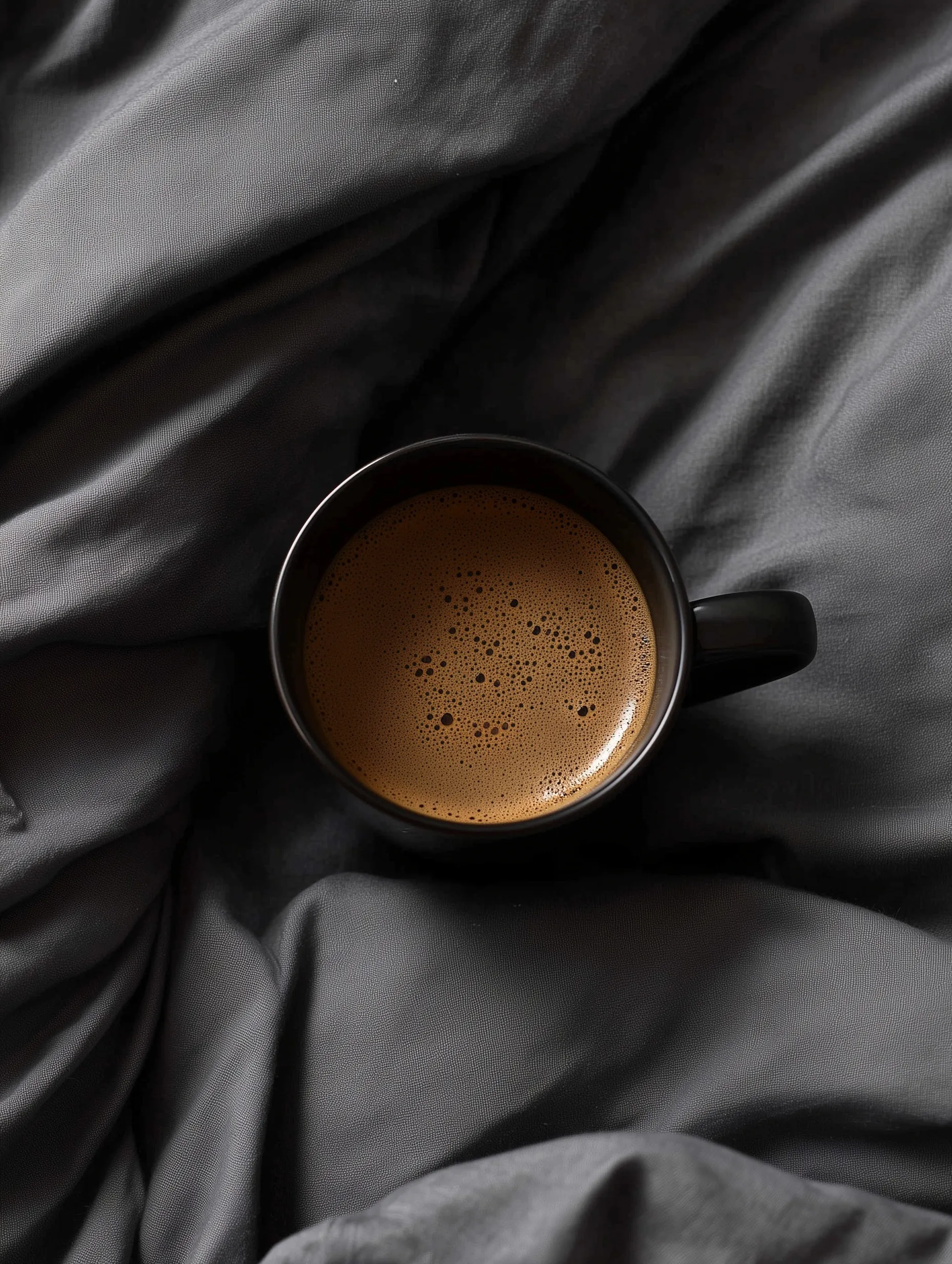 A steaming cup of black espresso on cozy, wrinkled dark gray bedding in a minimalist morning setting.