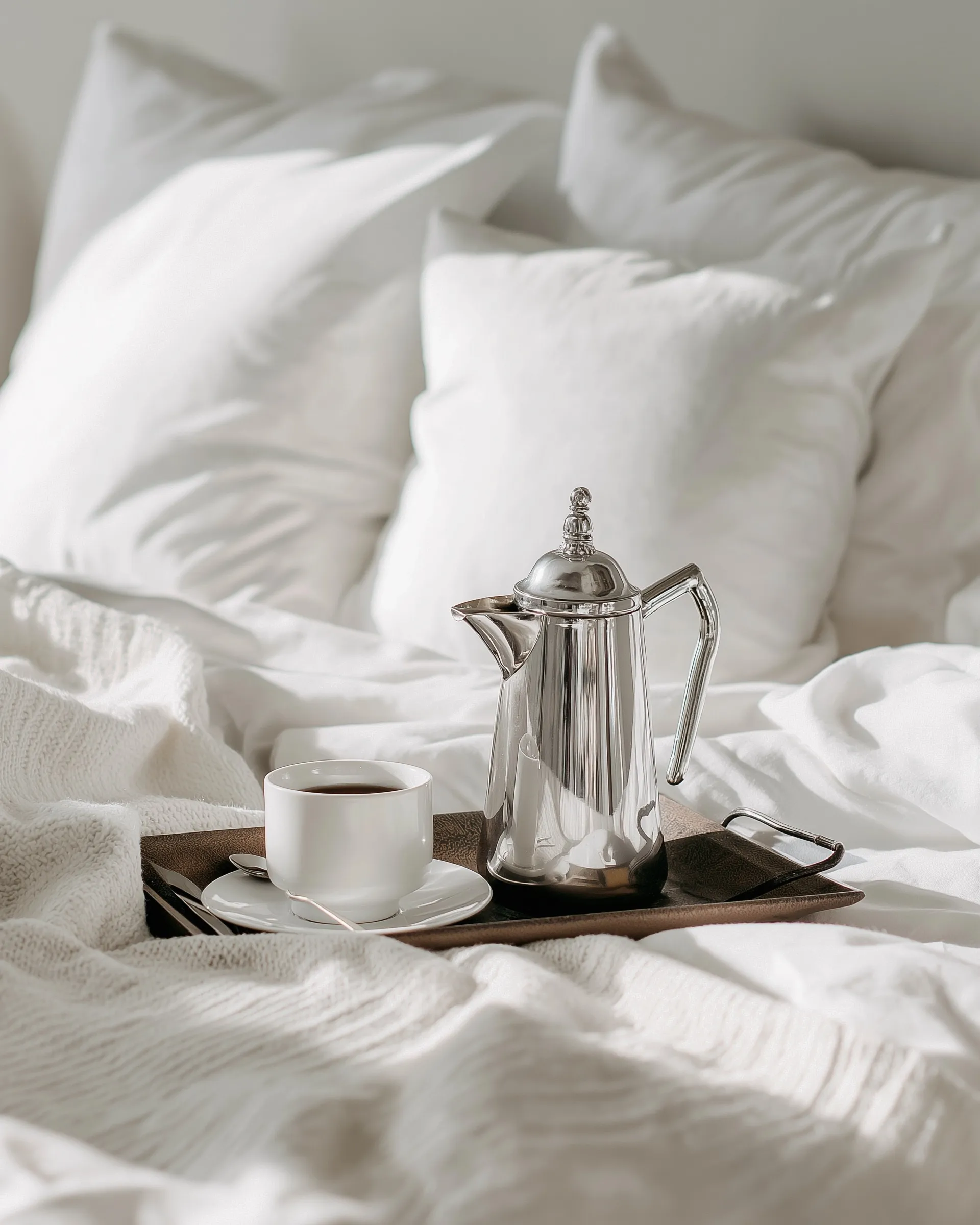 A silver coffee pot and cup on a tray over white bedding, creating a cozy morning aesthetic.