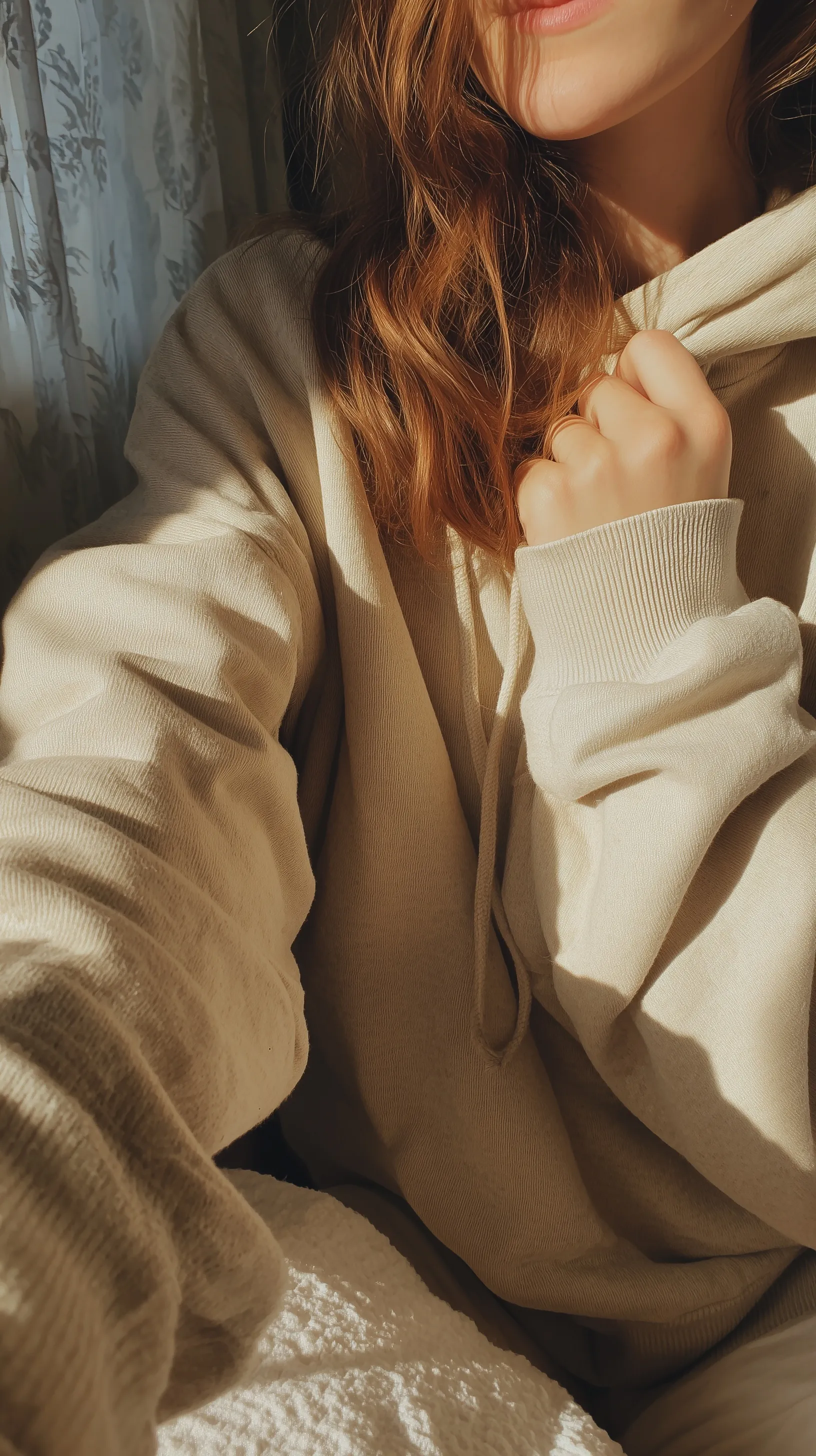 A young woman in an oversized beige sweatshirt taking a casual, candid handheld selfie in soft morning light.