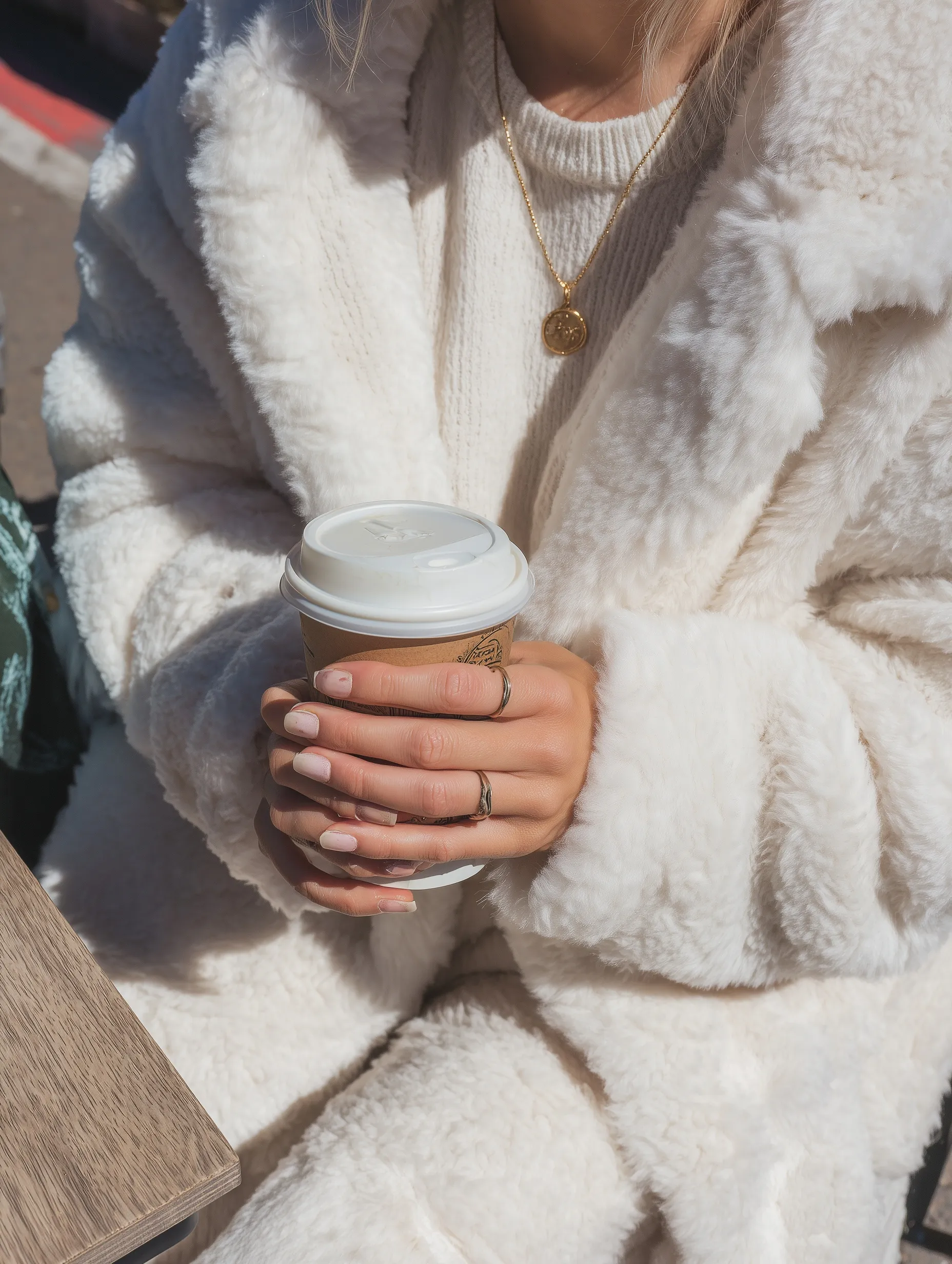 A stylish woman in an oversized white faux fur coat holding a coffee cup, captured in a sunny, vintage film style.