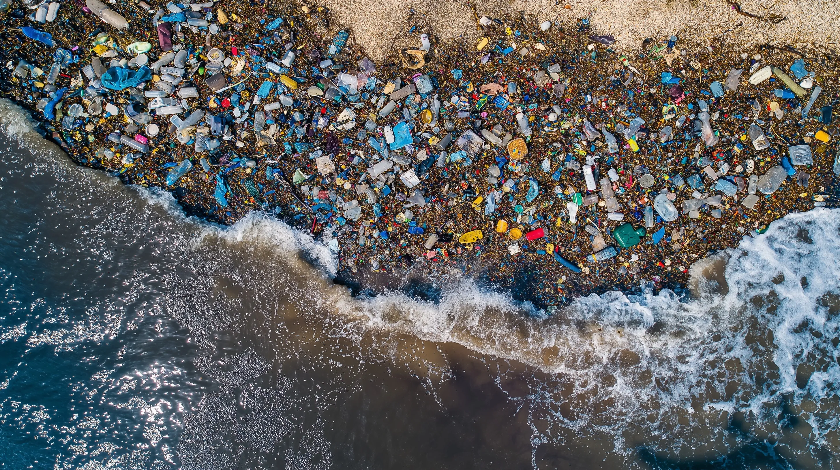 Birdseye view of excessive plastic bottles polluting ocean water along a sandy beach shoreline.