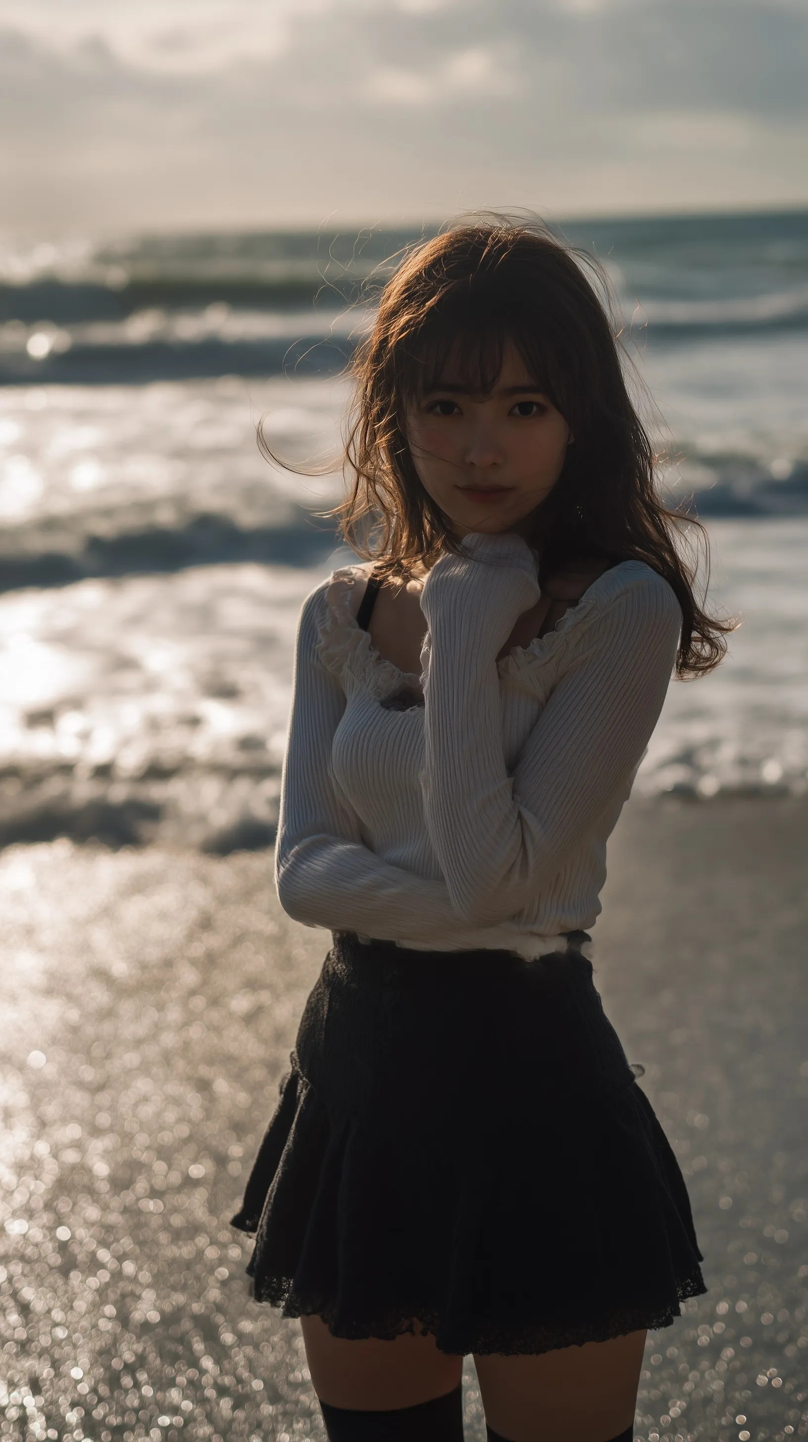 A shy Japanese girl in a white top and black miniskirt standing on a sunny beach with ocean waves.