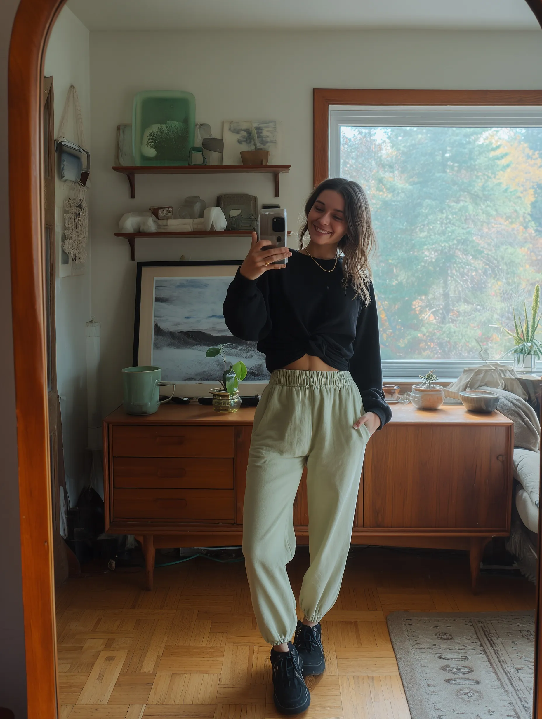 A woman in a black ribbed tank top taking a mirror selfie in a sunlit living room with wooden shelves.
