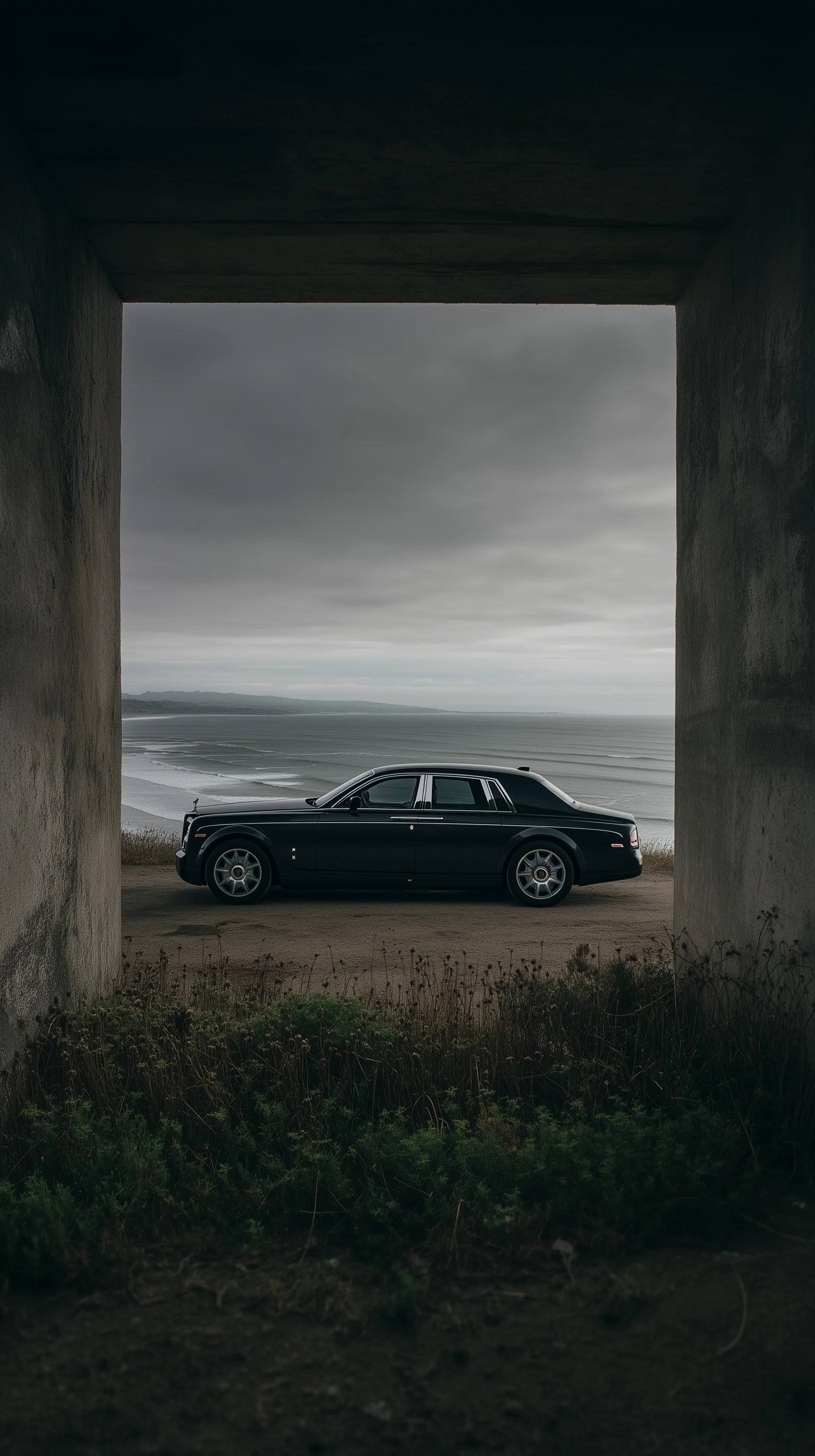 A sleek black Rolls-Royce Phantom parked in a concrete tunnel overlooking a moody coastal cliffside at twilight.