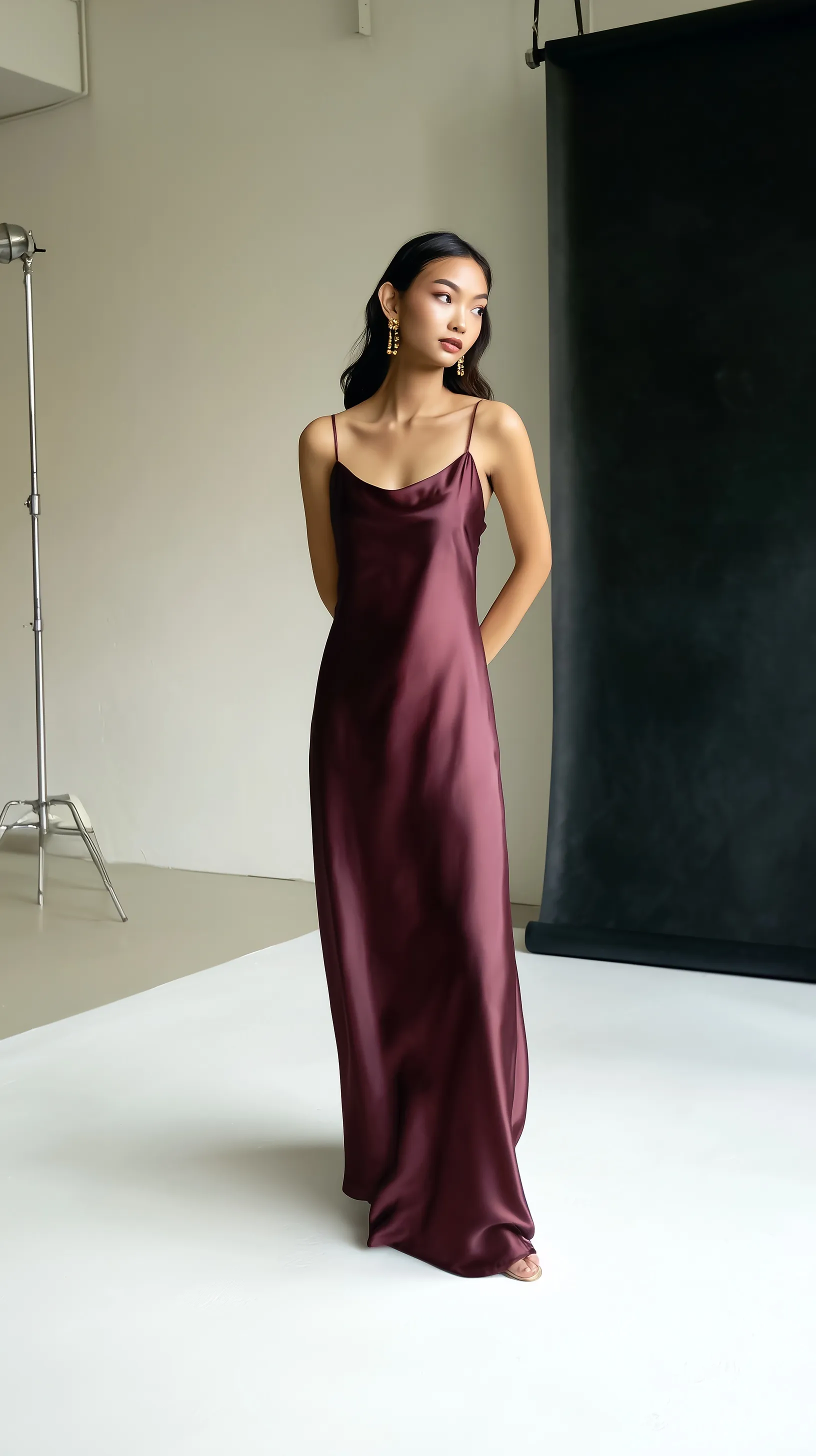 Elegant model in a deep-plum satin slip dress standing in a minimalist studio with soft natural lighting.