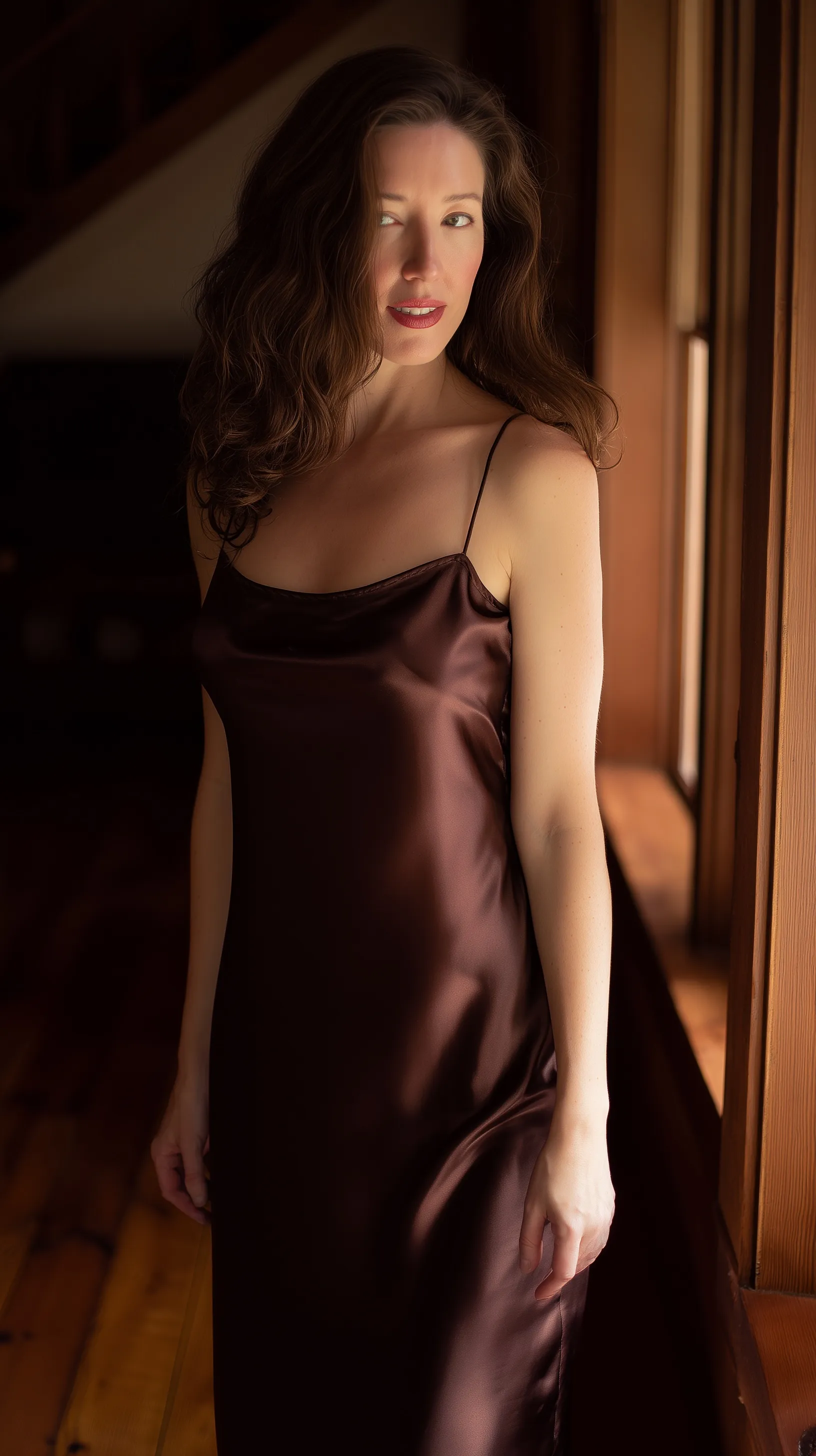 Elegant woman in a floor-length brown-burgundy silk satin slip dress standing in a luxury mansion, cinematic lighting.