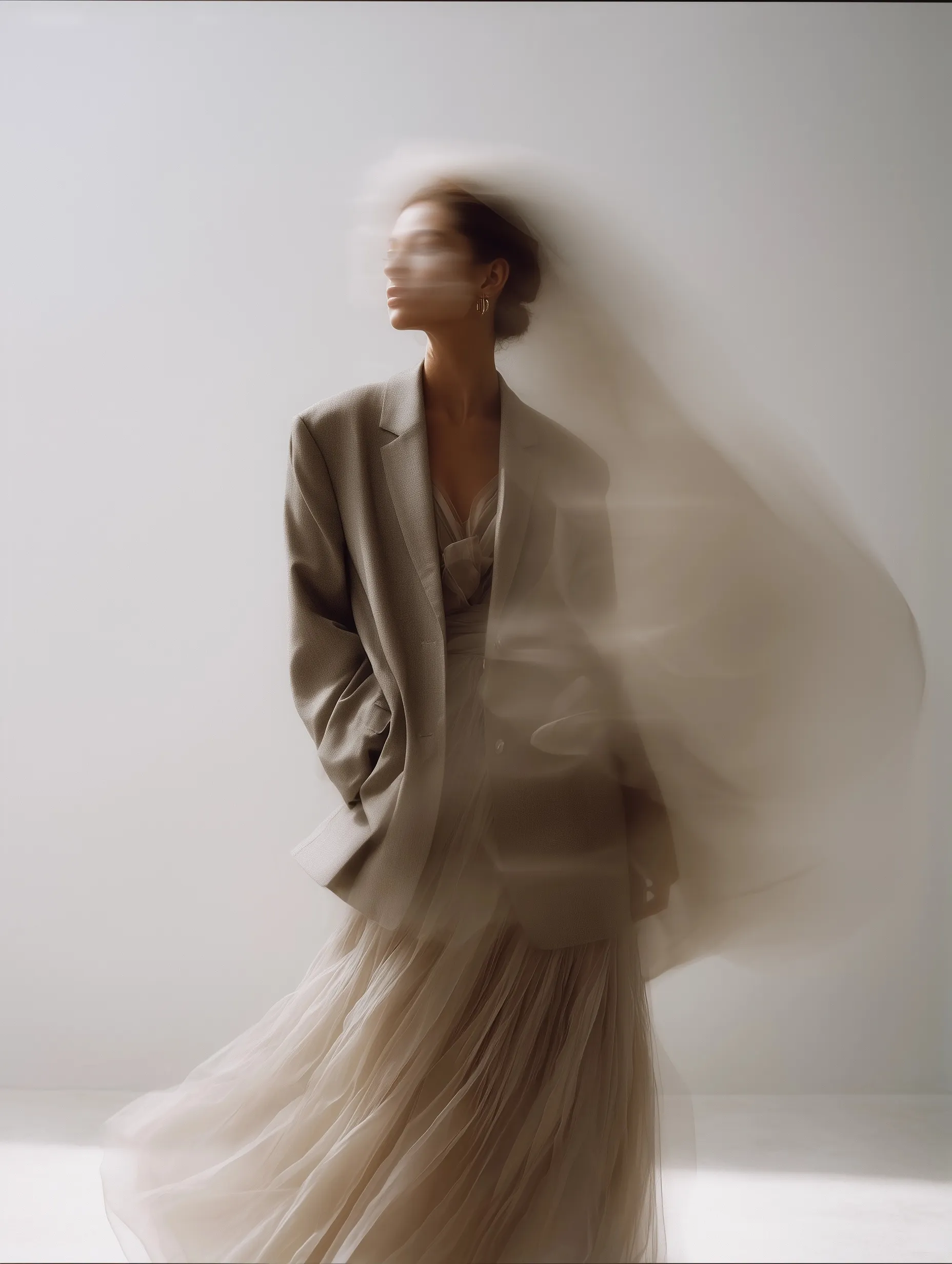 Fashion model in a taupe blazer and sheer beige pleated skirt posing in a minimal studio with flowing silk fabric.