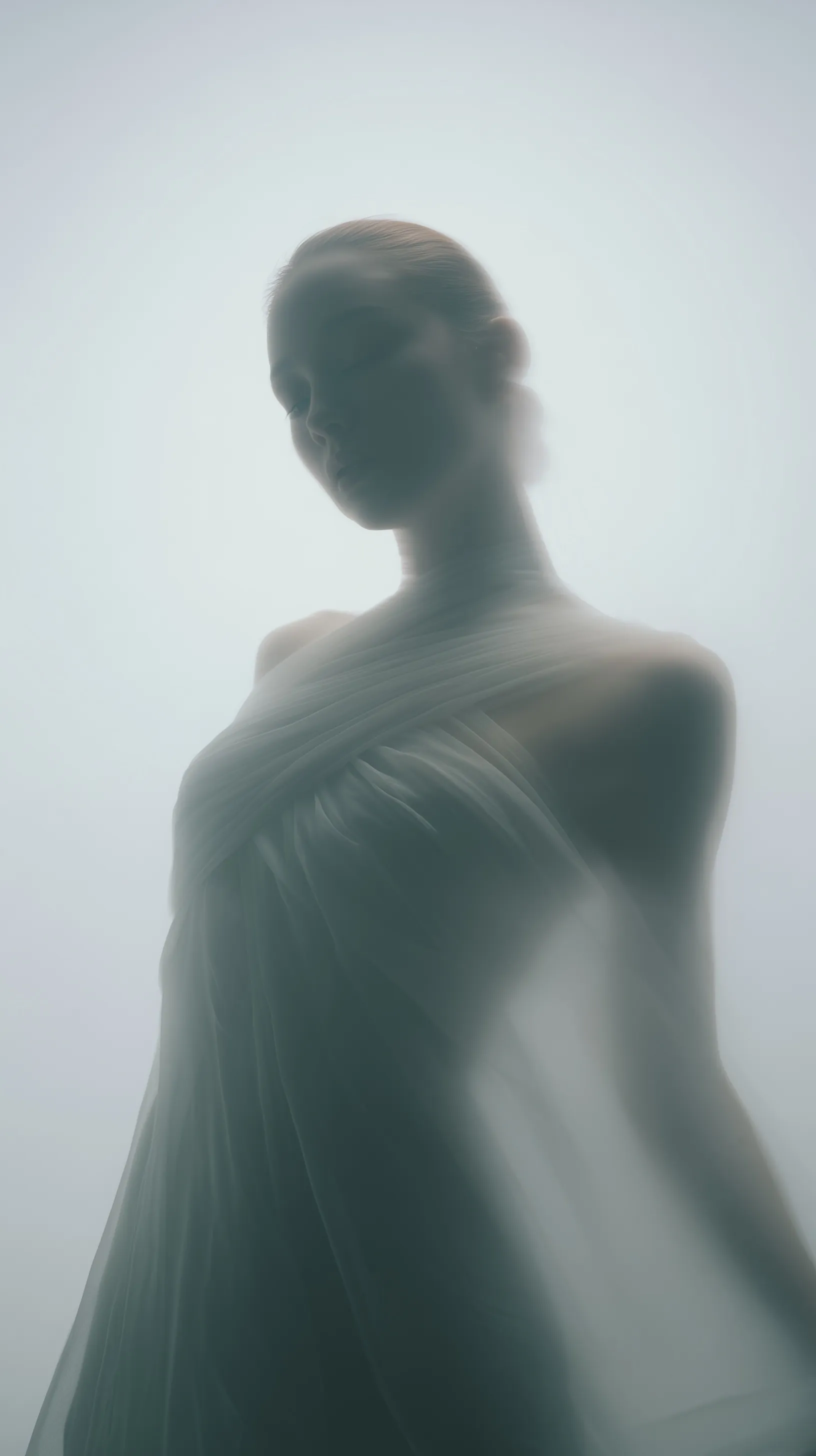Model in a sheer satin dress standing in a mist-filled studio with bright lighting and soft focus.