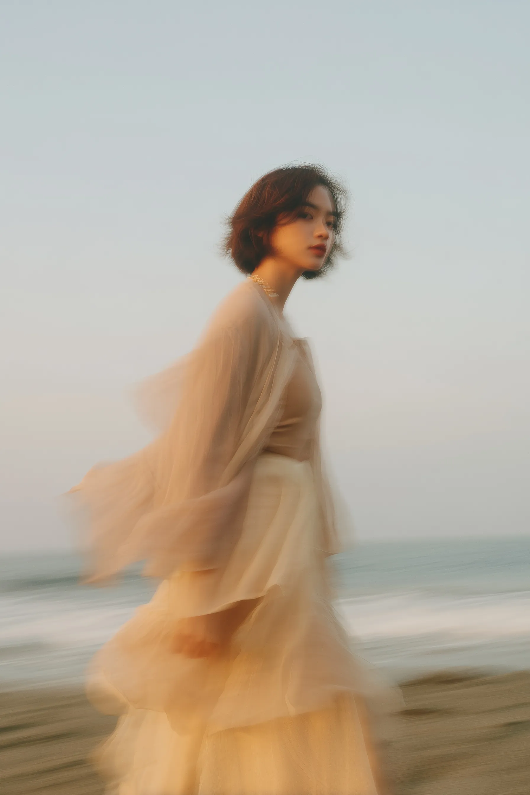 A young woman in a flowy, sheer beige layered dress walking on the beach with artistic motion blur.