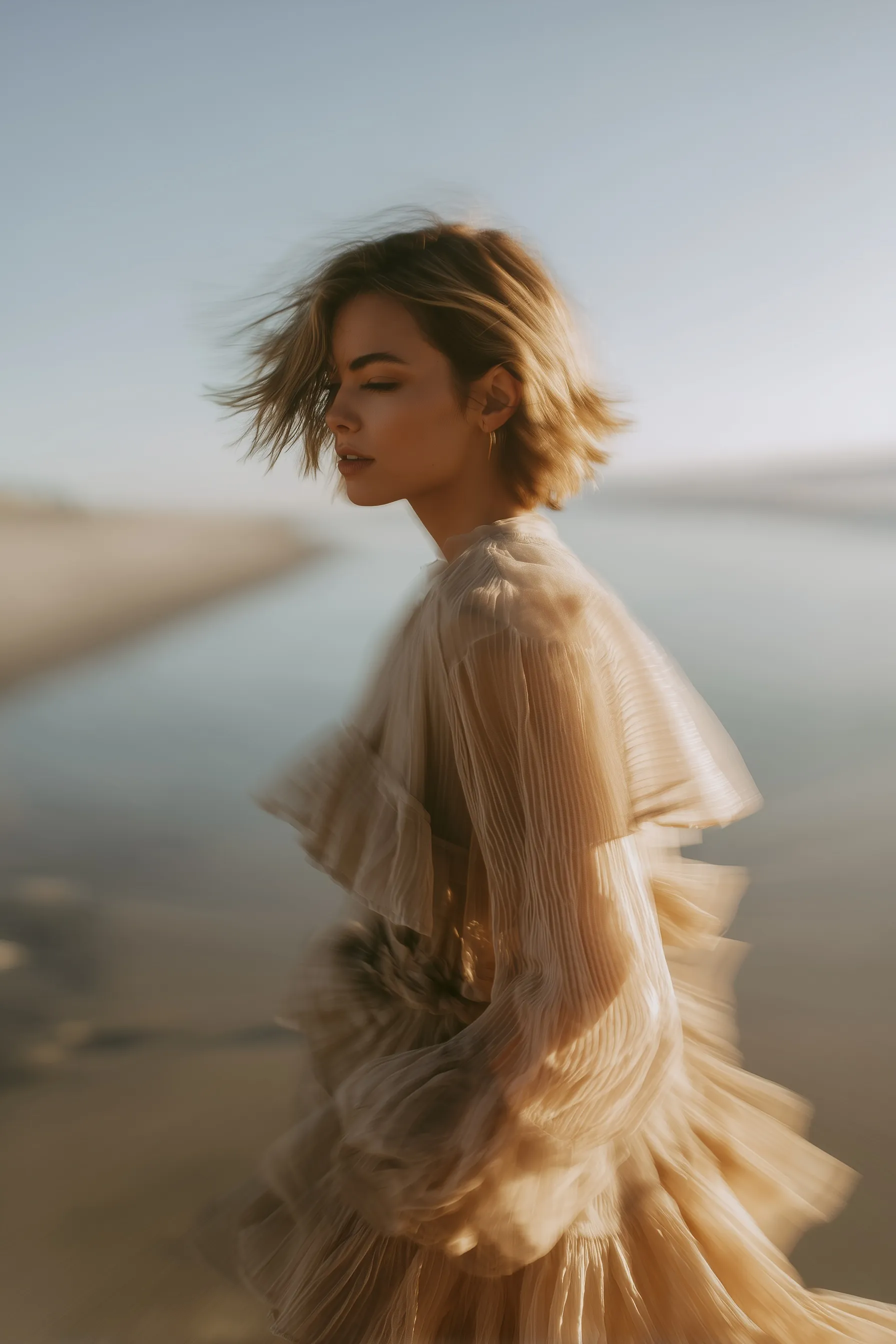 A young woman in a sheer layered beige dress walking along a beach with motion blur effect.