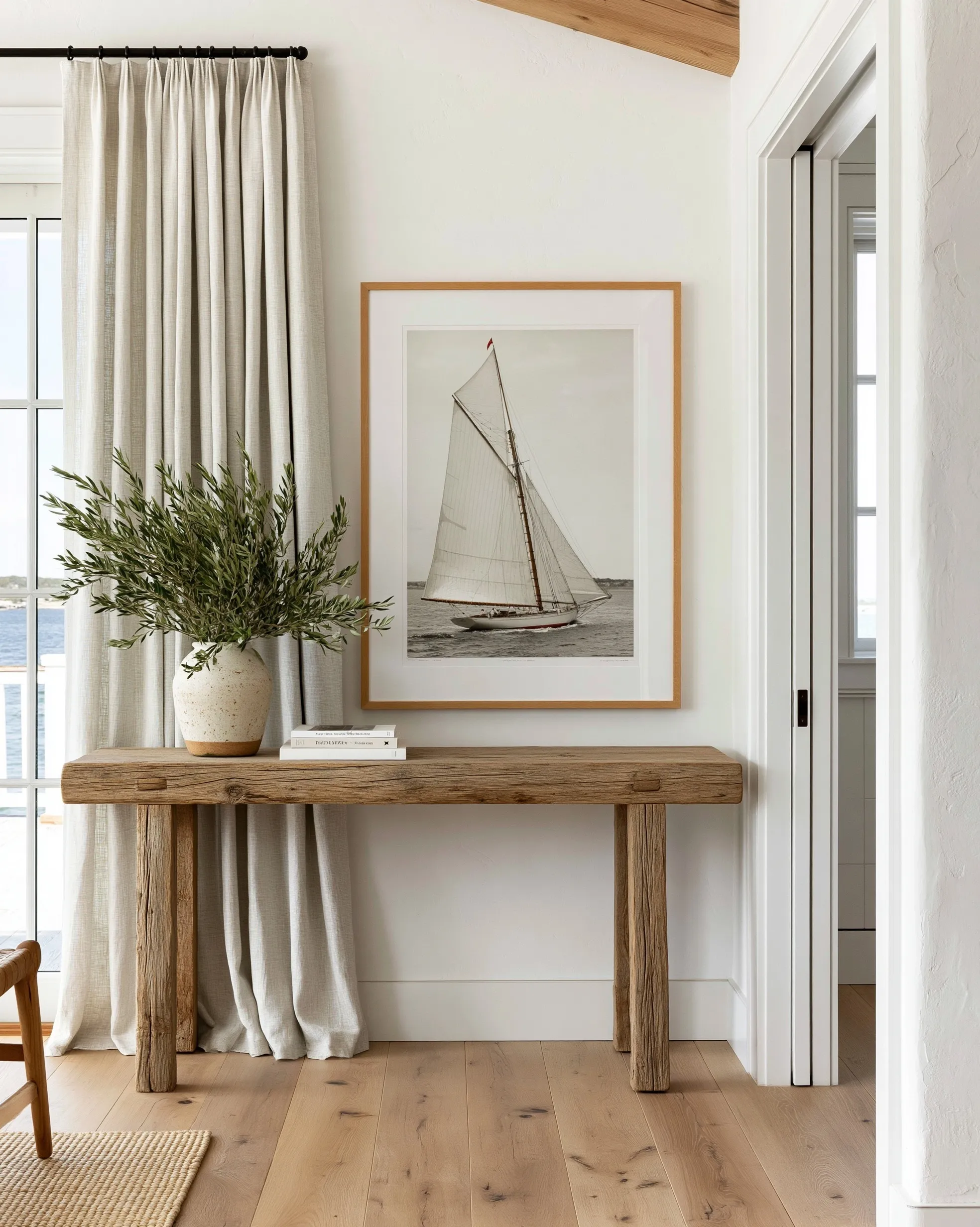 Vintage sailboat print above a rustic wood console table in a warm coastal cottage living room with olive branches.