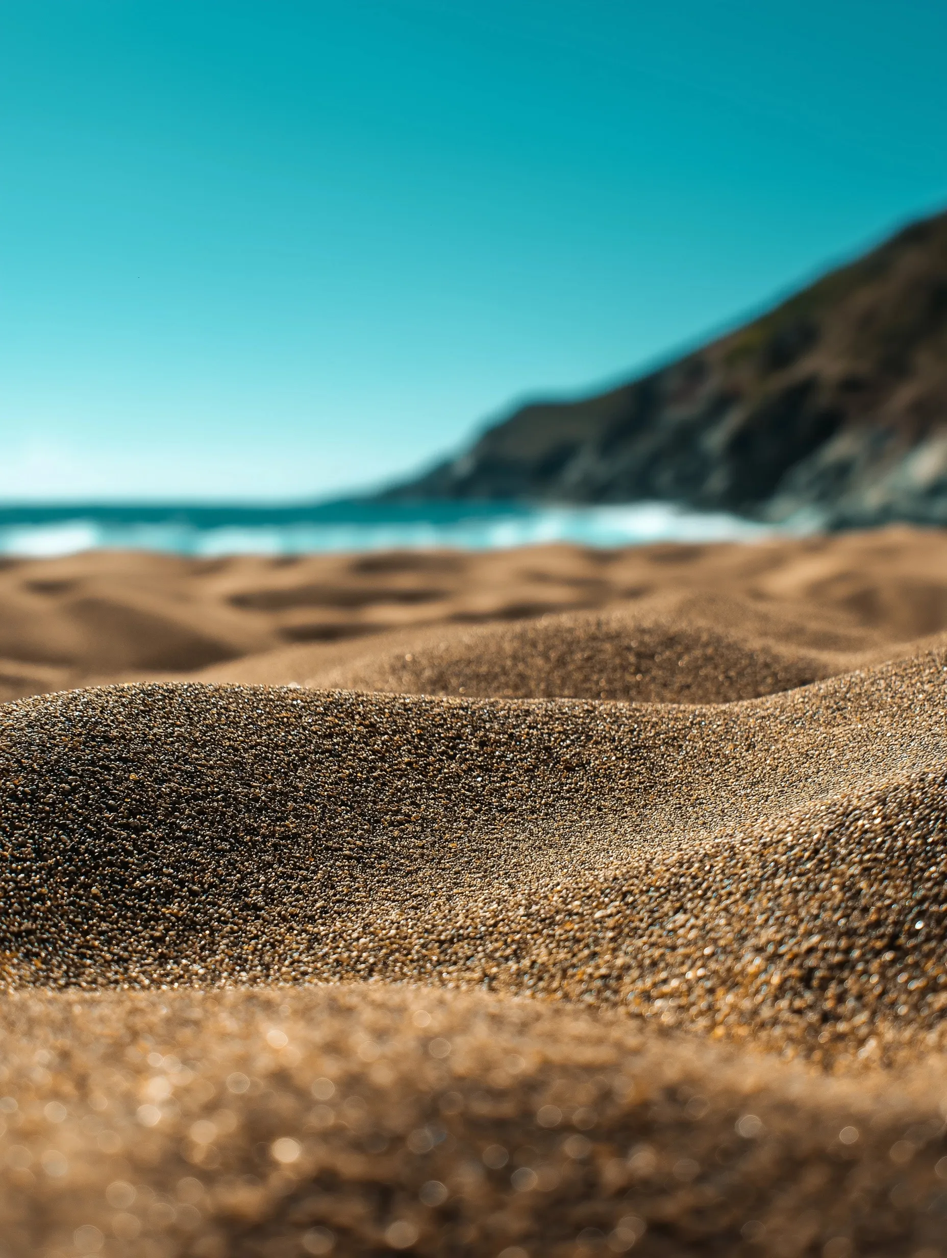 Close-up texture of golden beach sand with a soft blurred sea and clear summer sky background.