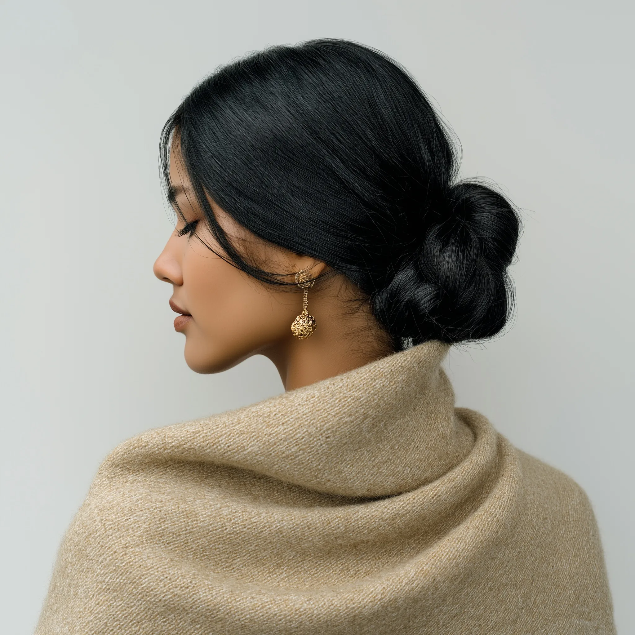 Indian woman with a sleek low bun hairstyle, wearing gold earrings and a wool coat, isolated on white background.