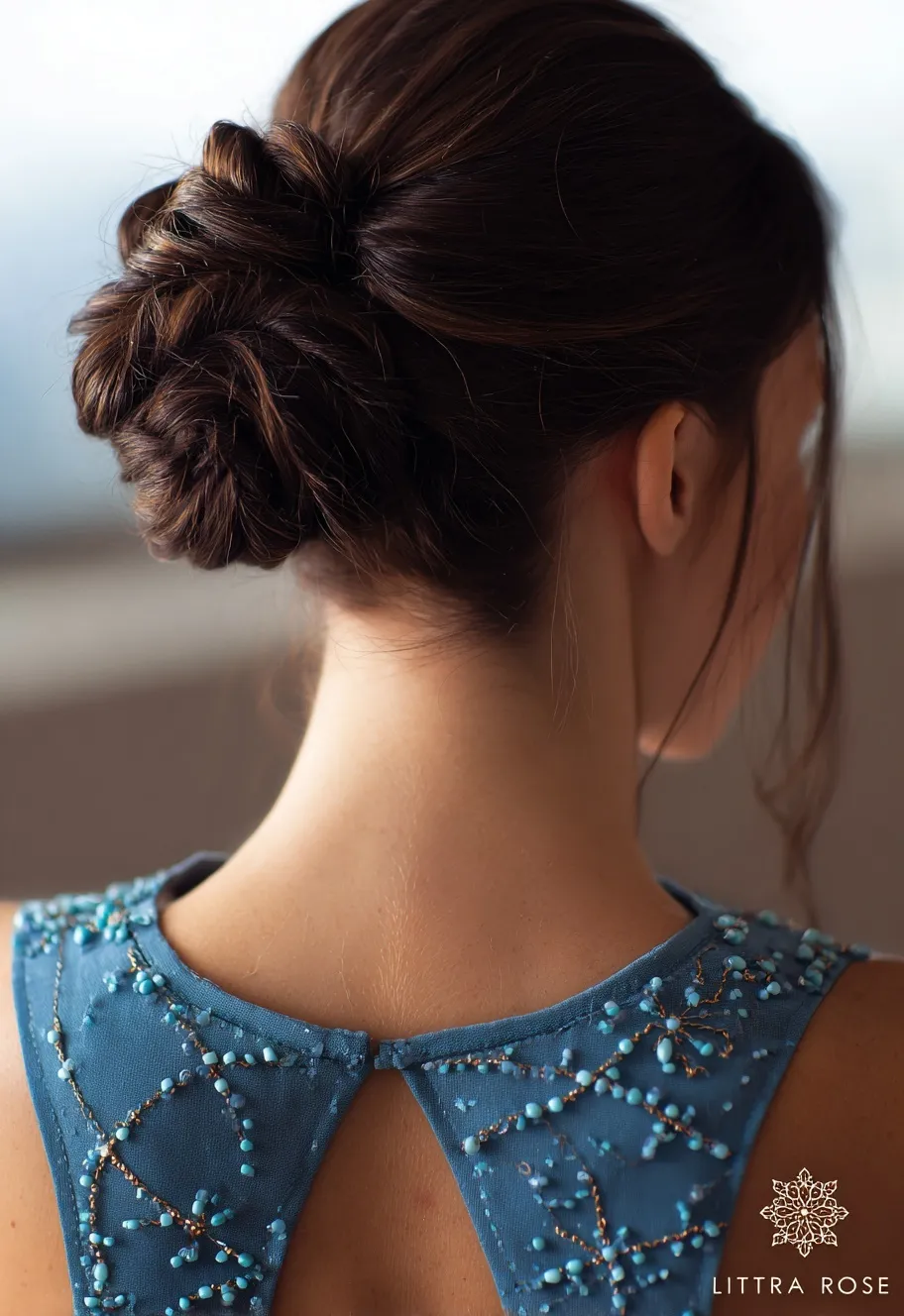 Close-up back view of a woman with an elegant dark brown wedding guest updo, styled in a bun with beaded blue top.