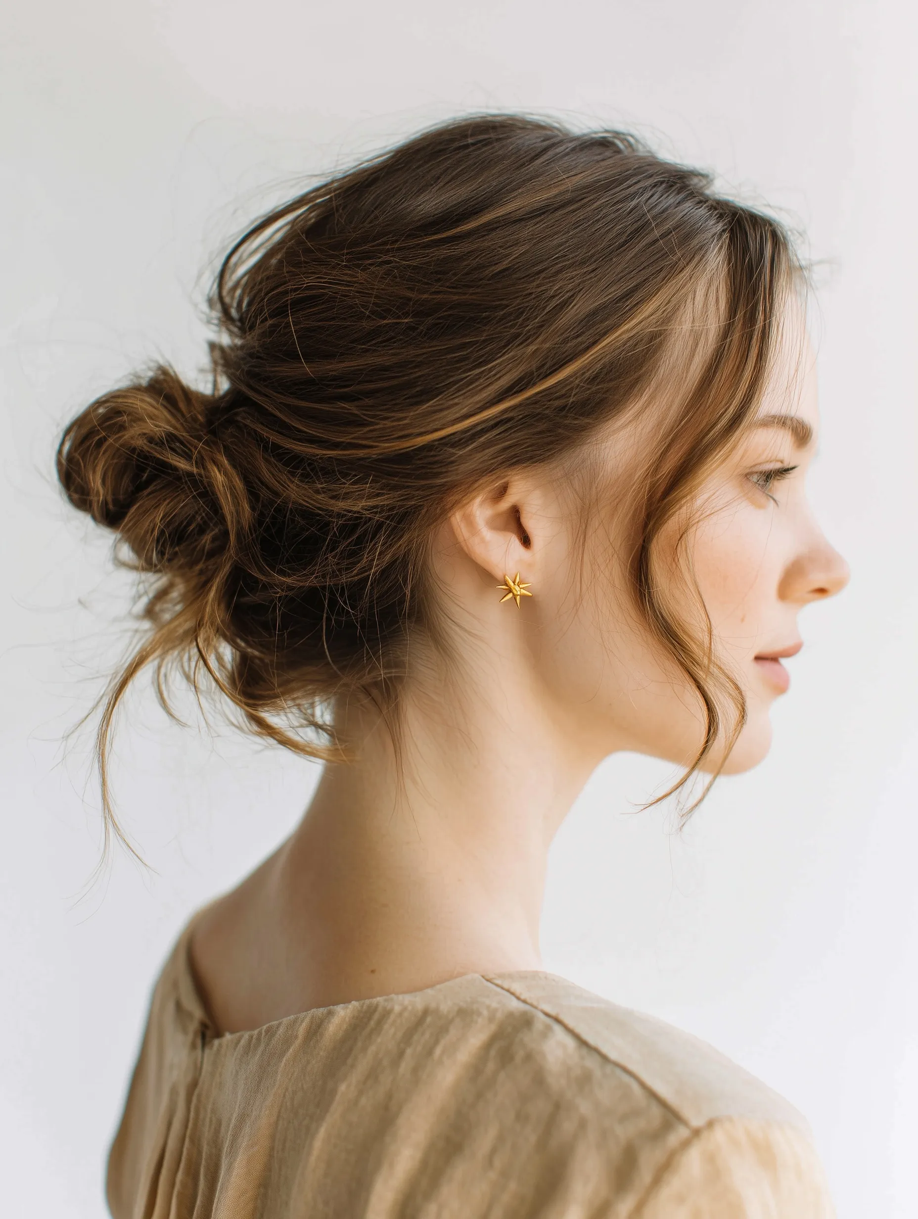Elegant wedding guest updo hairstyle with loose strands and gold star earrings, beige and white aesthetic.